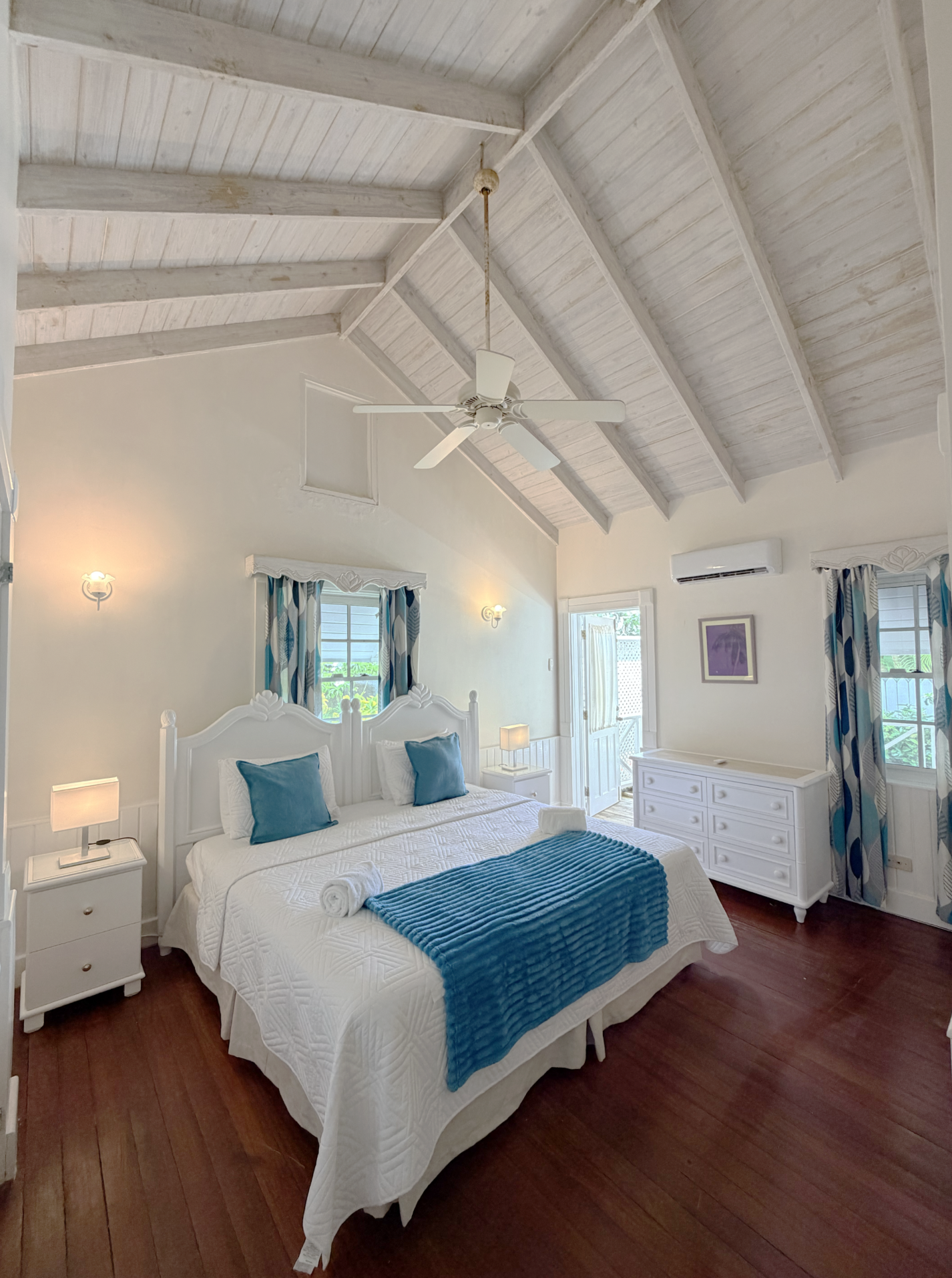 A bright bedroom with a white bed, blue pillows, and a blue blanket. There are two nightstands with lamps, and a white dresser. The room has high, vaulted, white-painted wooden ceilings with a ceiling fan and a mounted air conditioner. Two windows wi