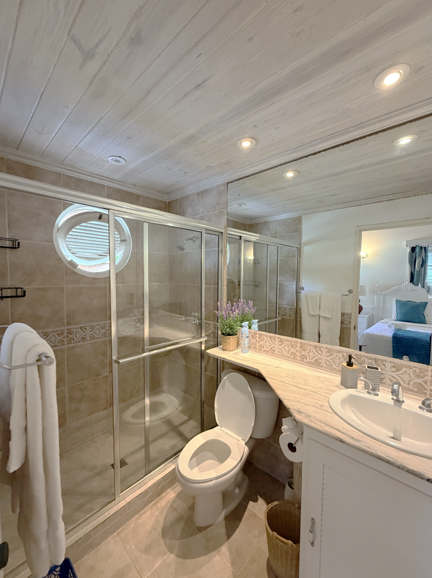 A bathroom with beige tiled walls and floor, a glass shower with metal frame, a small oval window, a toilet, a white vanity with a sink, a mirror, and various toiletries on the countertop. There is a towel rack with white towels, a basket on the floo