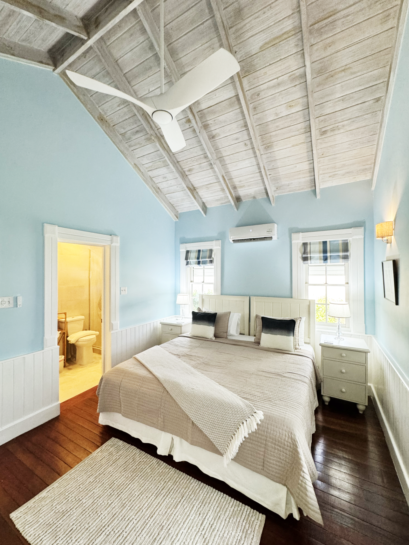A bedroom with light blue walls, white wooden wainscoting, and a vaulted ceiling with exposed wood beams. There is a ceiling fan with three blades. The bed has a beige quilt and four pillows, with a beige throw blanket. There are two white nightstand