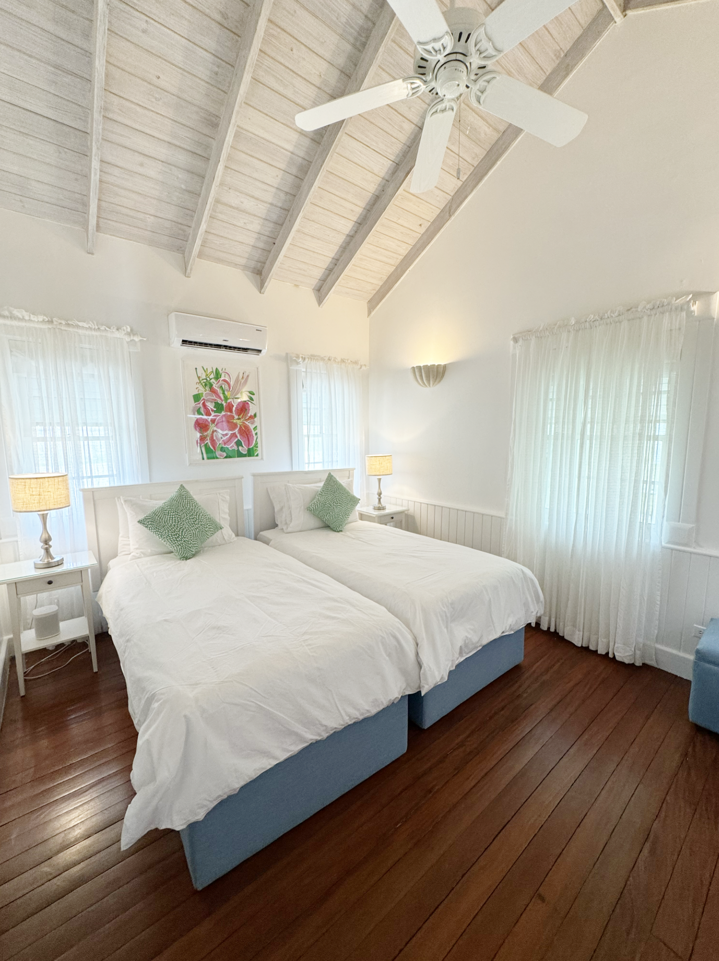 A bedroom with two beds, each with white bedding and green pillows. White nightstands on each side with lamps. Light-colored wood ceiling with exposed beams, a ceiling fan, and sheer curtains on windows. Artwork of pink flowers on the wall, wooden fl