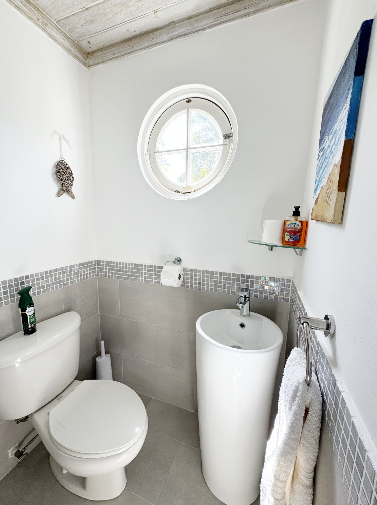 A small bathroom with a white toilet, a white bidet, and a round window with a view of palm trees outside. There is a towel hanging on a rail, a shelf with toilet paper and hand soap, and decorative wall accents.
