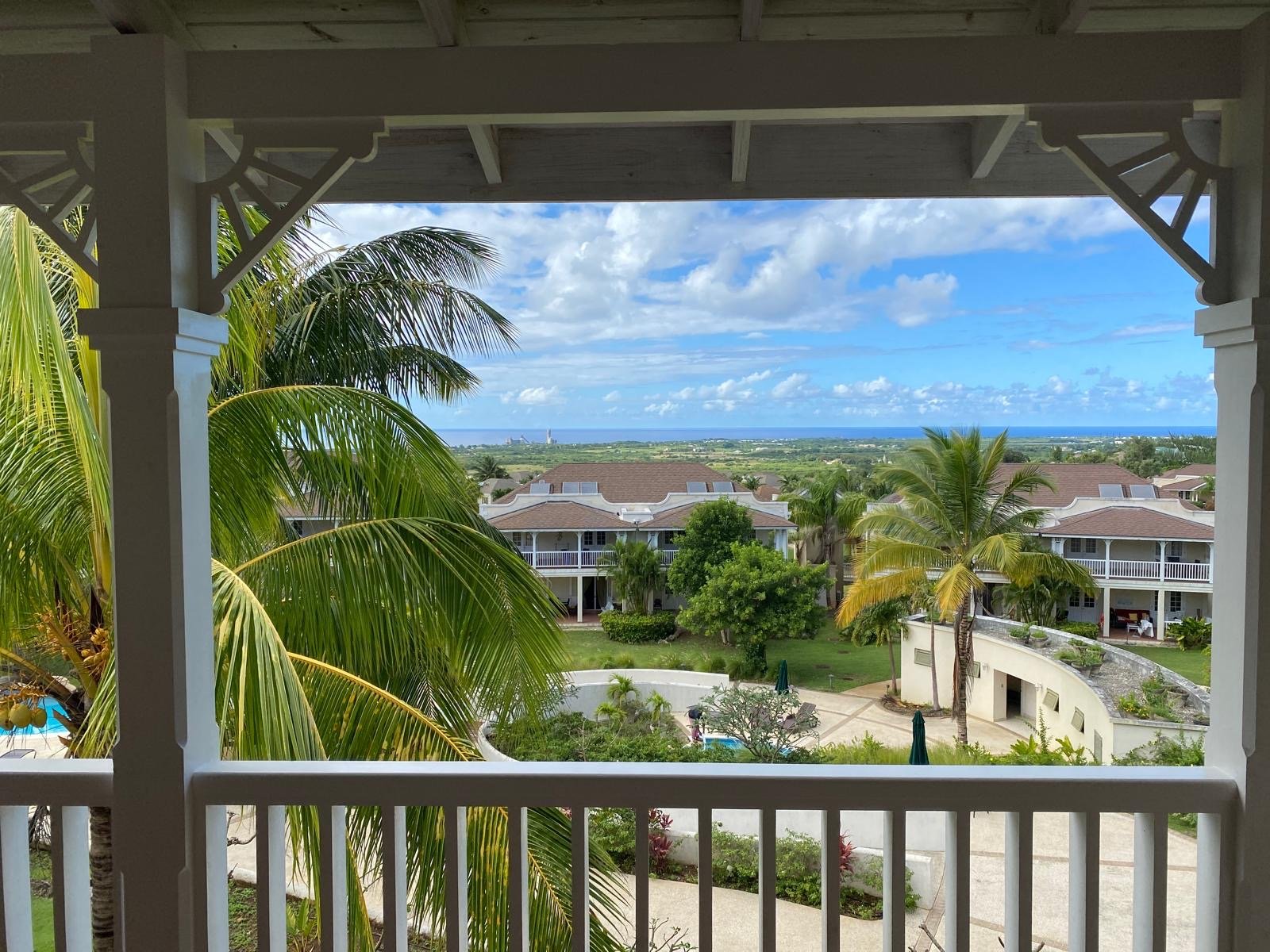 View from a balcony showcasing tropical palm trees, residential buildings with balconies, a courtyard with a swimming pool, and a distant ocean under a partly cloudy sky.