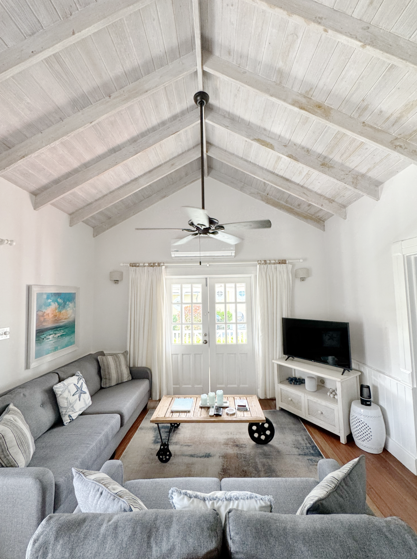 Living room with a vaulted ceiling, ceiling fan, white curtains, and a door with glass panes, shelves with decor, and a sofa with decorative pillows.