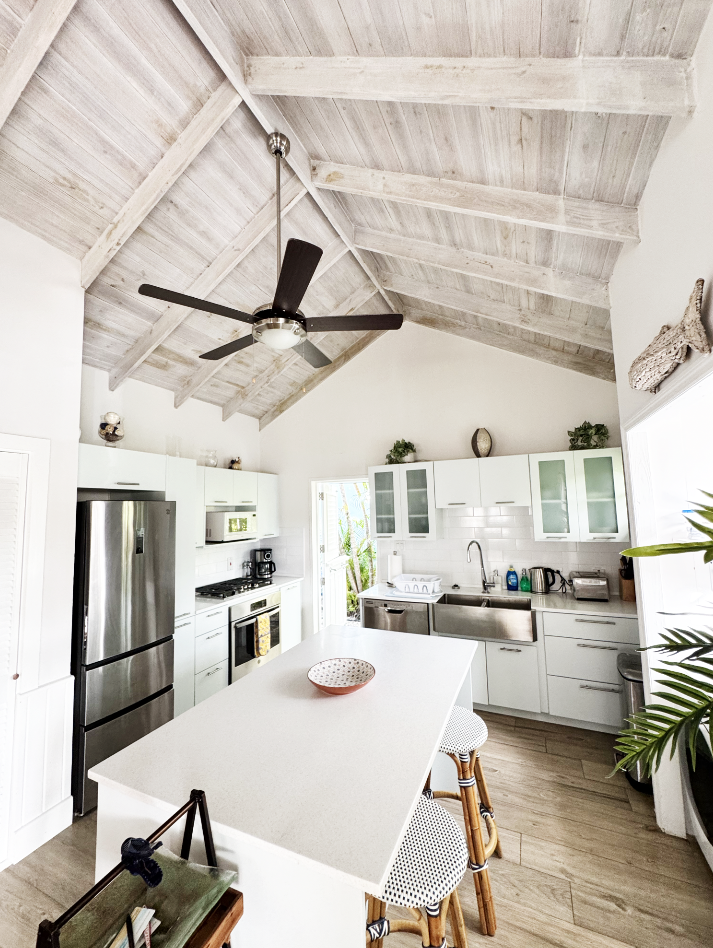 A modern kitchen with white cabinets, stainless steel appliances, and a high, wood-paneled ceiling with a ceiling fan. There is a white island in the center with seating, and decorative objects on top of the cabinets.