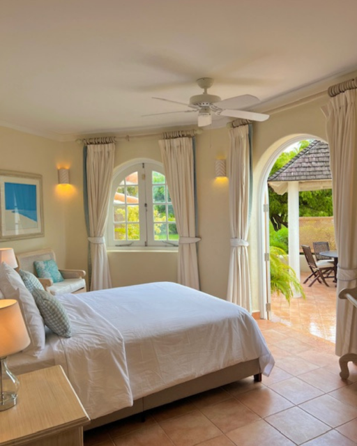 A bright bedroom with a large bed, white bedding, and multiple pillows, near a window with white curtains, leading to an outdoor patio with a table and chairs, surrounded by lush greenery.