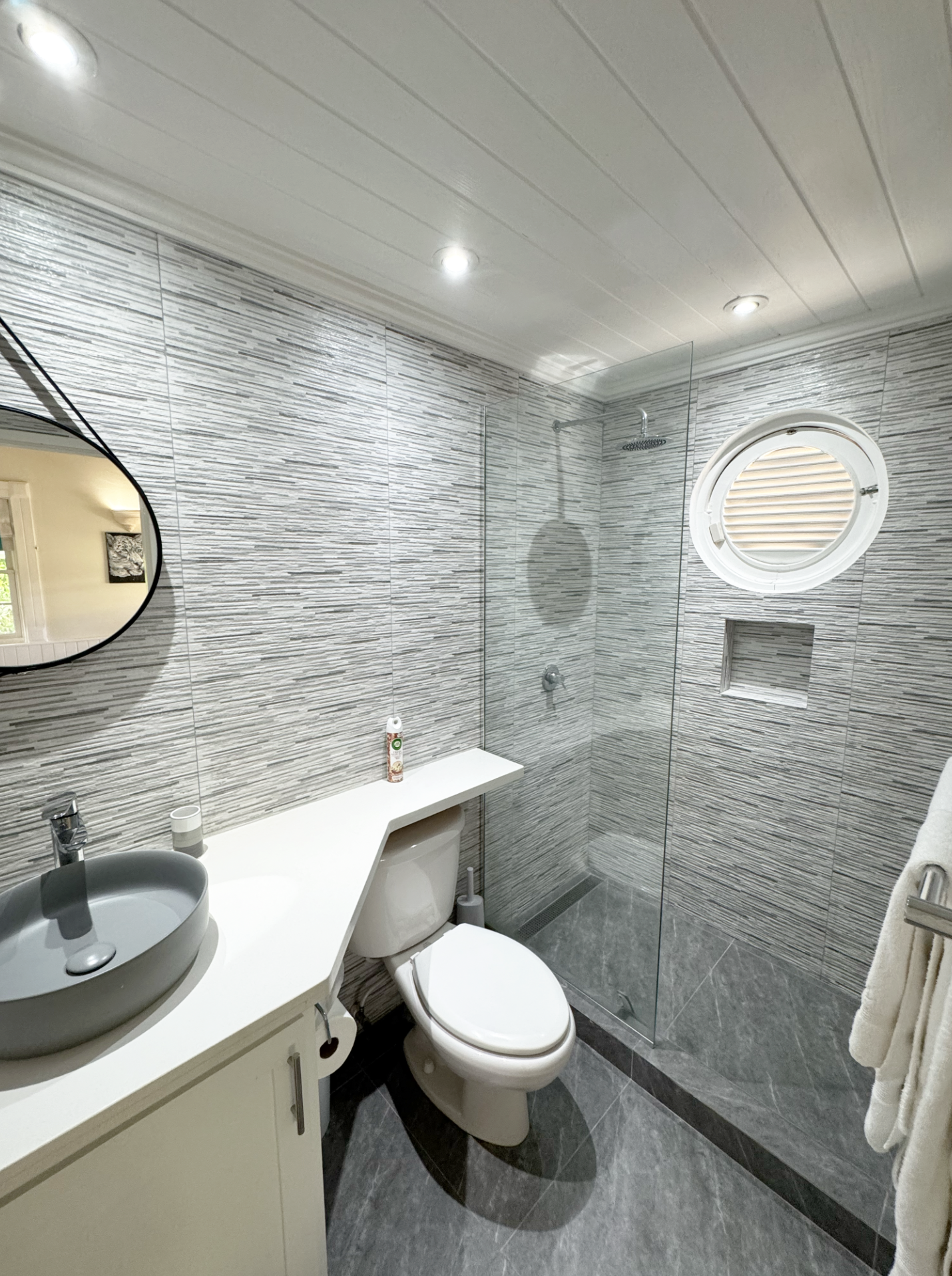A modern bathroom with a grey tiled wall, a small round mirror, a white vanity with a vessel sink, a toilet, and a walk-in shower with a small window, a showerhead, and a glass partition.