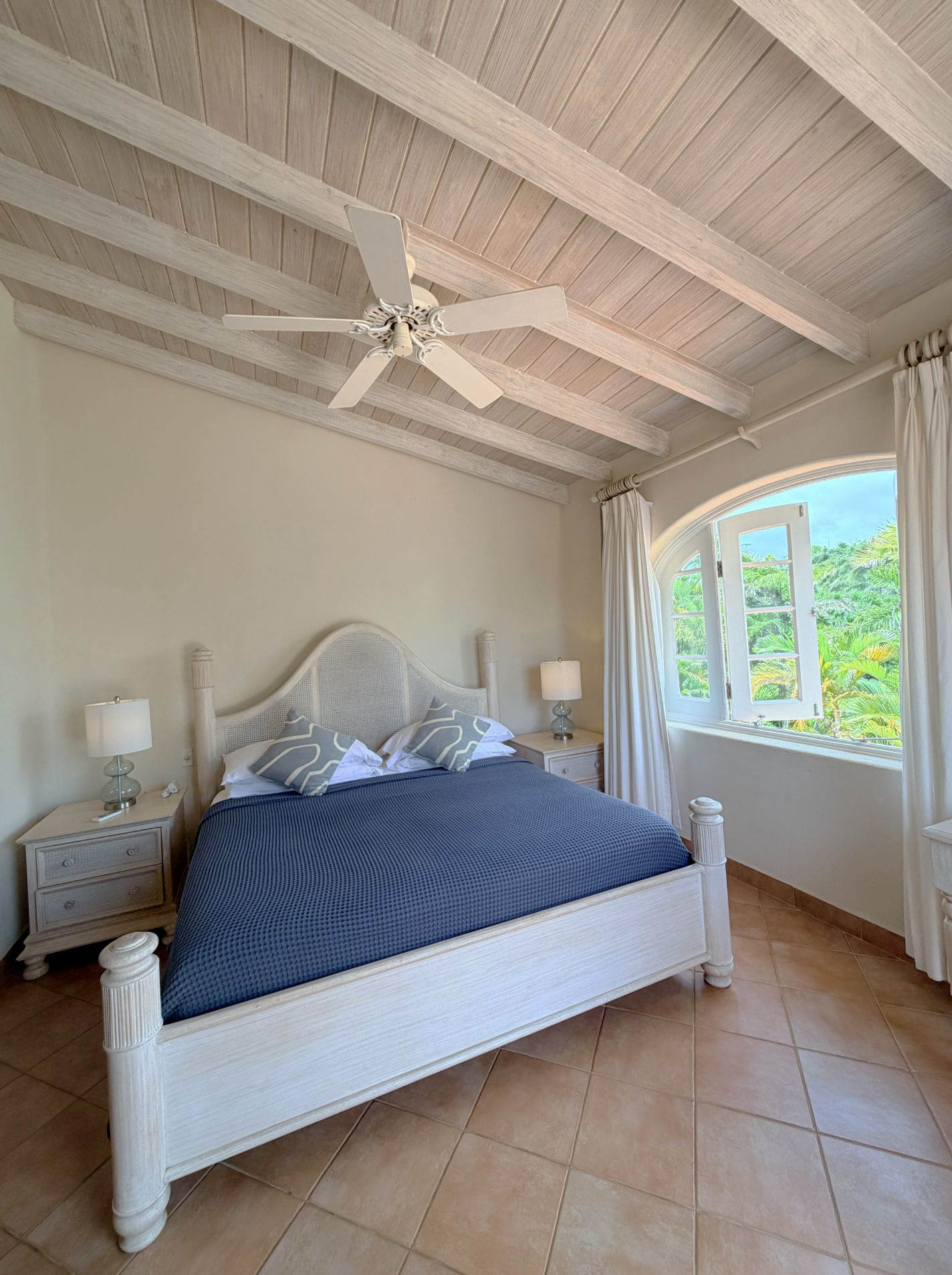 A cozy bedroom with a white wooden bed, blue bedding, two matching pillows, two bedside tables with lamps, a window with white curtains, and a ceiling with exposed wooden beams and a white ceiling fan.