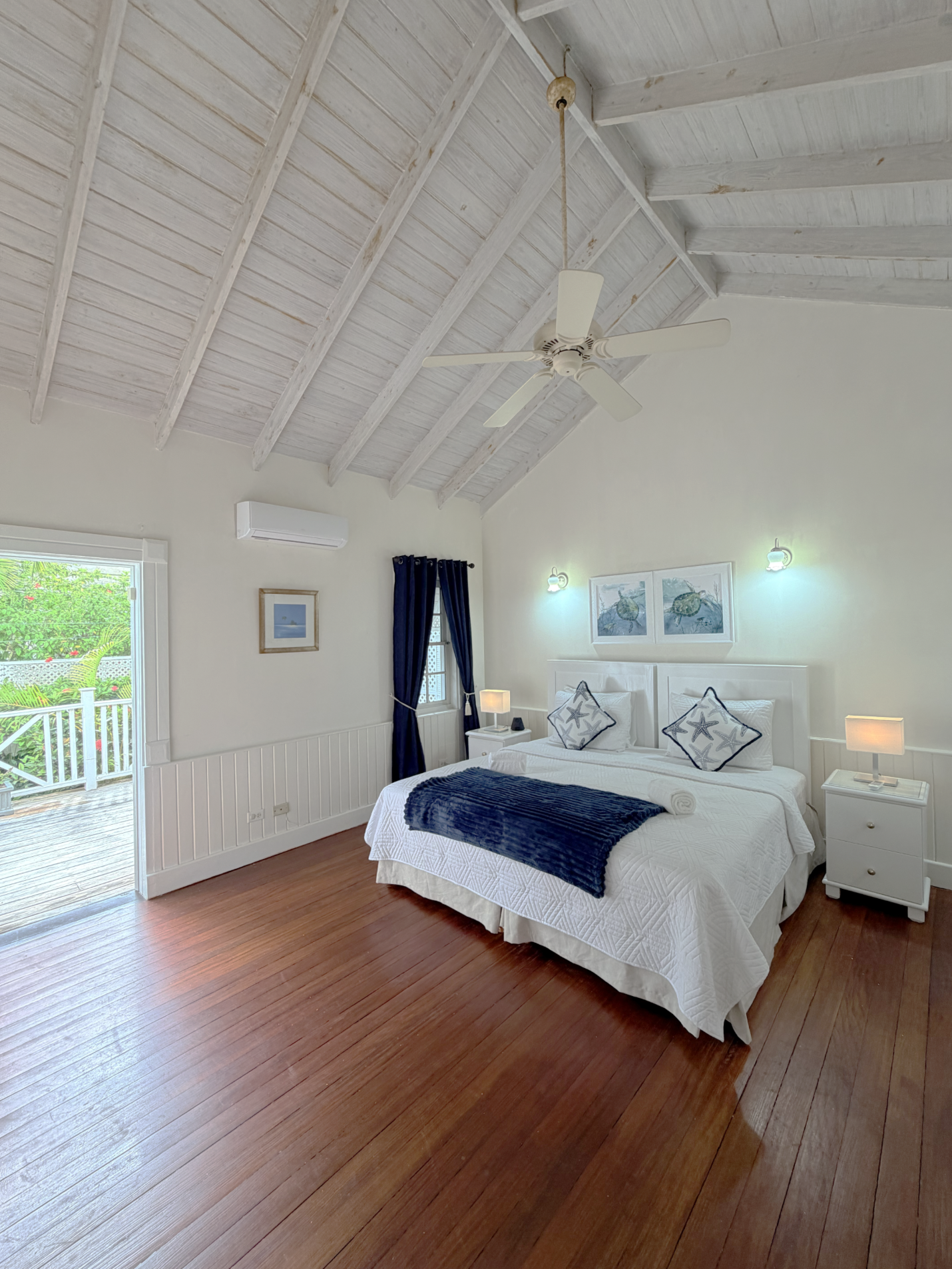 A bedroom with a white bed, navy blue throw blanket, decorative pillows, white nightstands with lamps, wall art, dark blue curtains, a portion of a patio with greenery outside, a ceiling fan, and wood flooring.