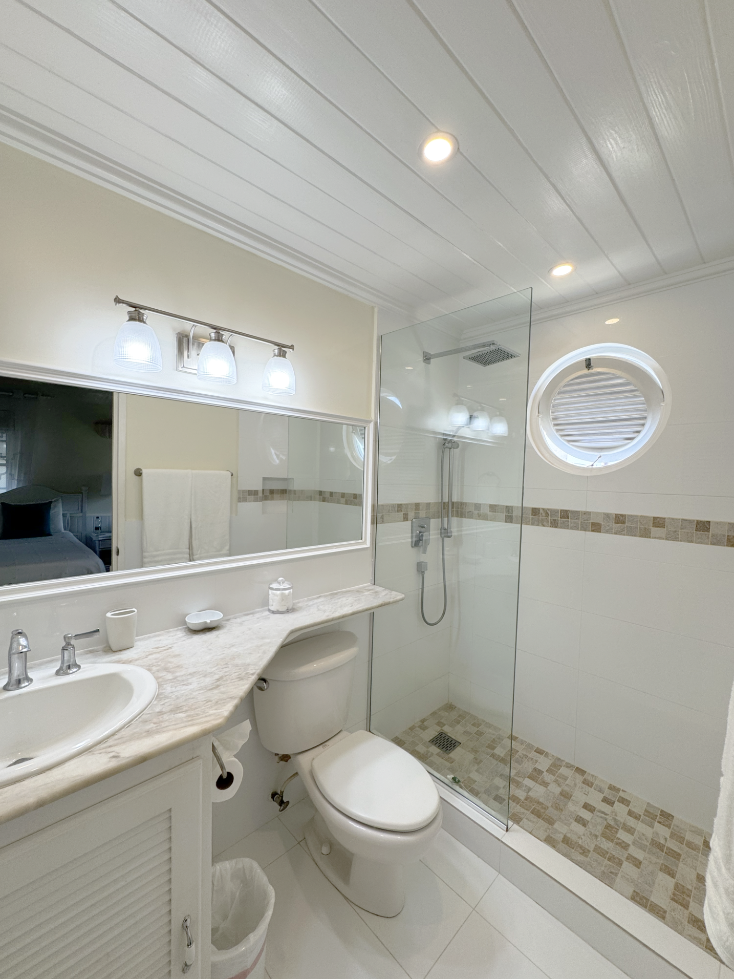 A modern bathroom with a white vanity, marble countertop, and a toilet. There is a glass shower enclosure with a rain showerhead, a circular frosted window, and a mirror above the sink. The ceiling has recessed lighting and a light fixture with three