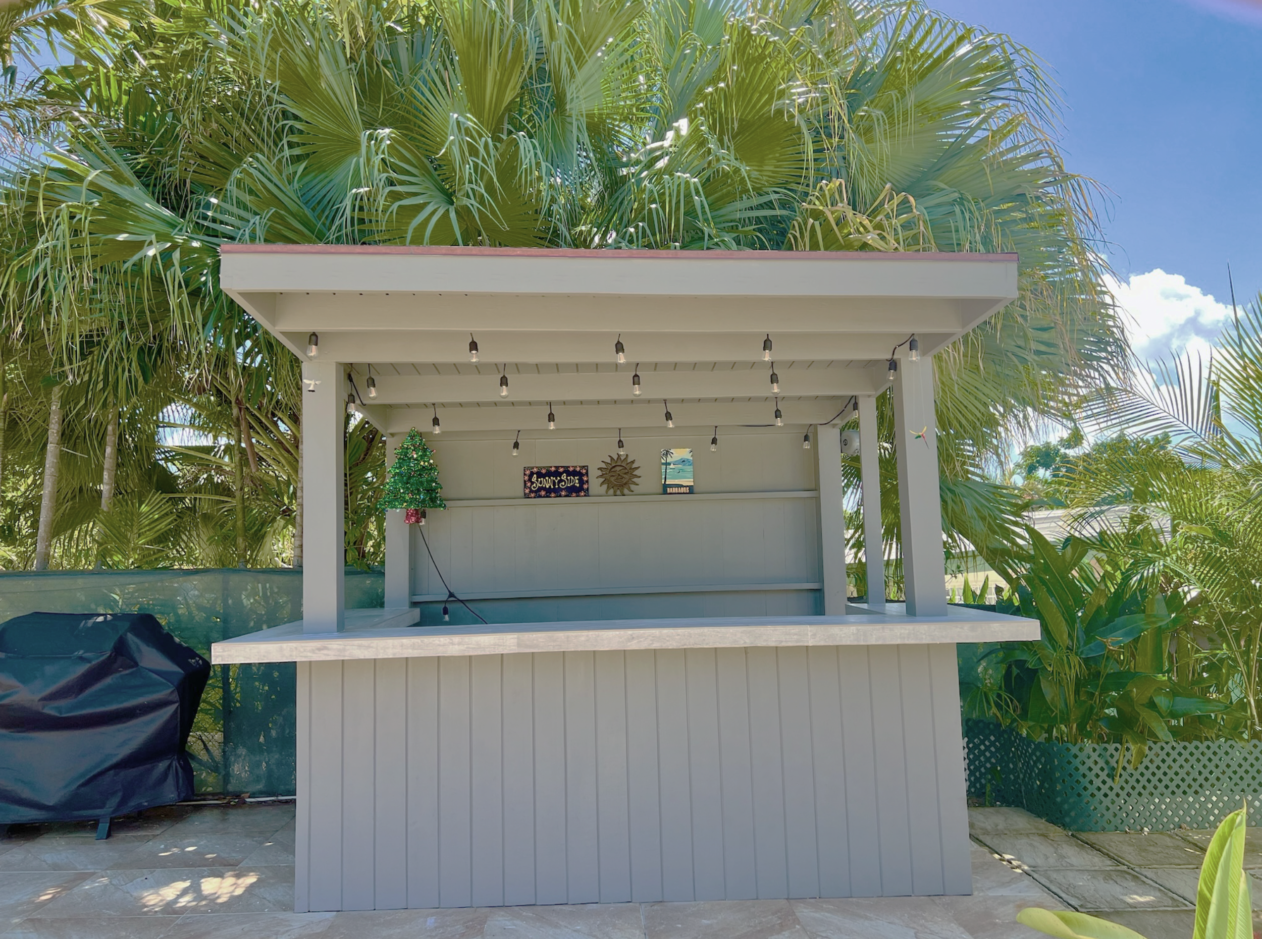 A white outdoor bar or counter with string lights hanging from the roof, decorated with a small Christmas tree, set against lush green palm trees and a partly cloudy blue sky.