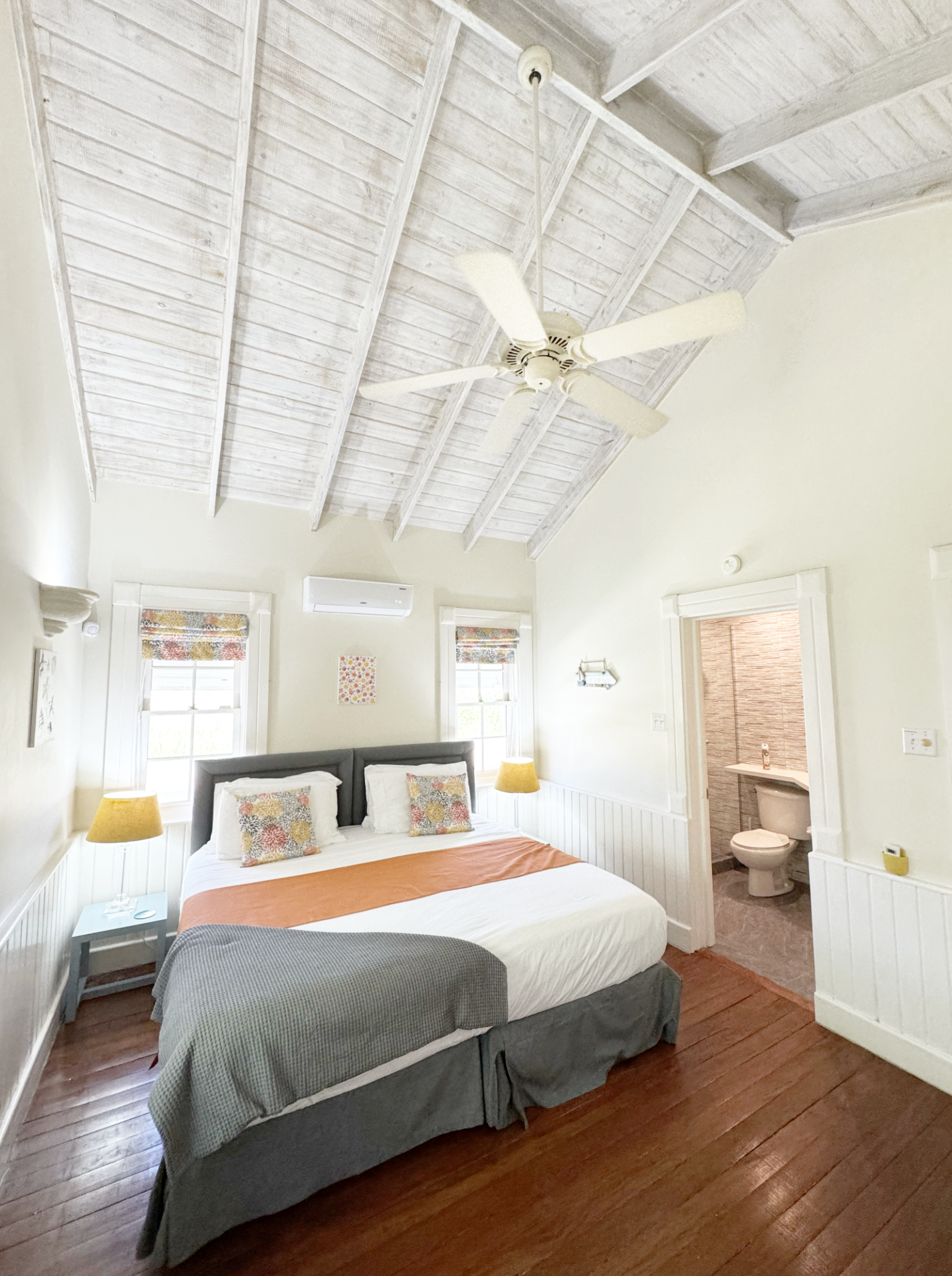 A bedroom with a bed, colorful pillows, nightstands with yellow lamps, hardwood floors, windows with curtains, and a ceiling fan. An adjacent bathroom is visible through an open door.