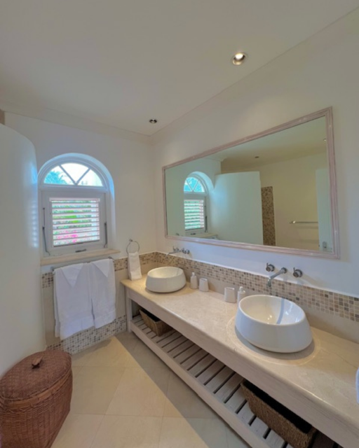 A bathroom with a large mirror, two vessel sinks, a window with shutters, and decorative tiles, featuring a wicker laundry basket in the corner.