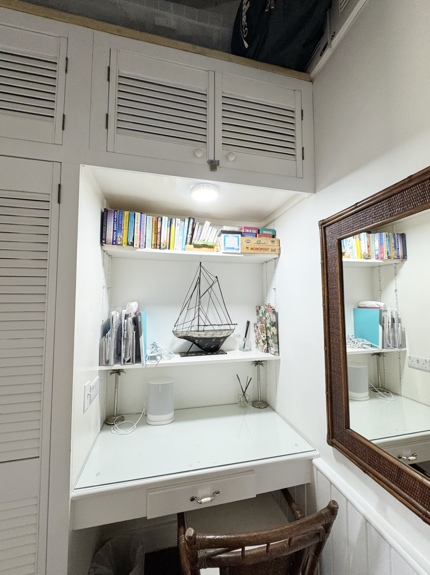 A white desk with a glass top, decorated with a nautical boat model, a lamp, and some office supplies. A mirror with a wooden frame is on the right side. The background features a bookshelf with books and a box, along with a white wall and a ceiling 