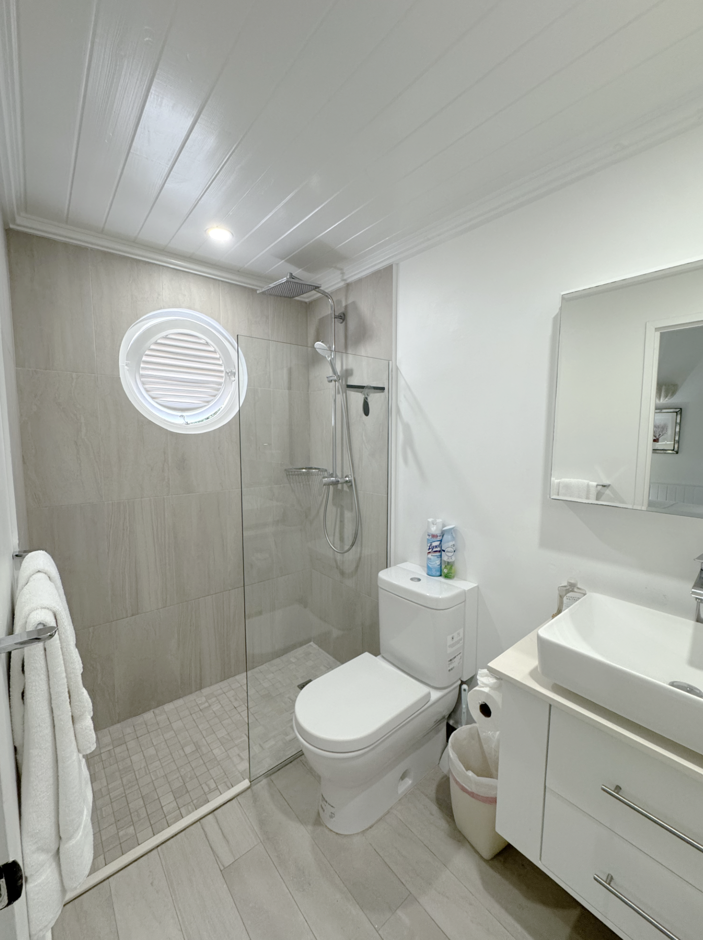 A modern bathroom with a walk-in shower enclosed by clear glass, a round window with shutters, a toilet, a white vanity with a sink, a mirror, and supplies on the countertop.