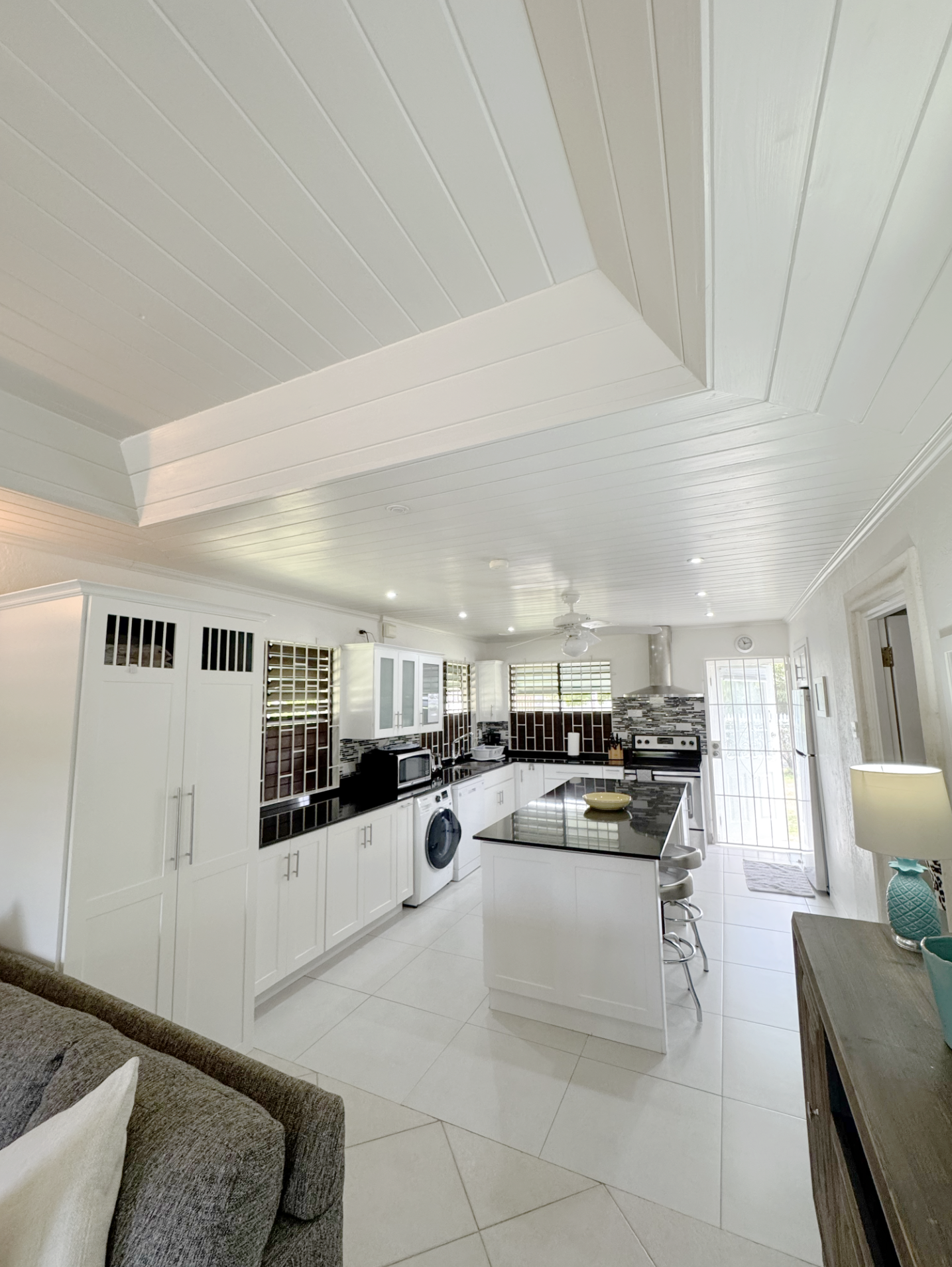 Bright kitchen with white cabinets, black countertops, and tiled backsplash. Ceiling has beadboard paneling and a ceiling fan. Appliances include a microwave, washing machine, and oven. Tall cabinet and bar stools at kitchen island. Large windows wit
