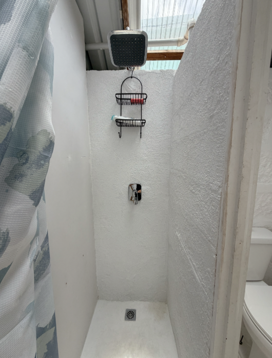 Shower stall with a black showerhead attached to the ceiling, two wall-mounted wire racks for toiletries, a white textured wall, and a small drain on the shower floor.