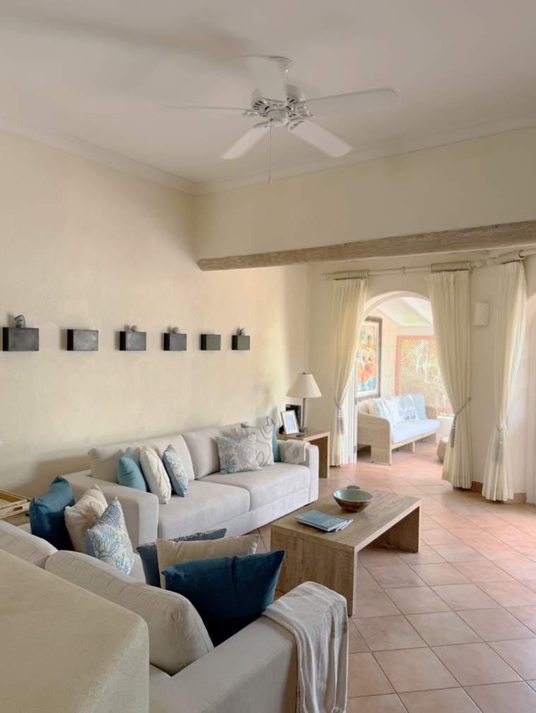 Living room with cream-colored sofas, blue and patterned pillows, a wooden coffee table, a side table with a lamp, a ceiling fan, and an arched doorway with curtains leading to a sunlit patio with additional seating.