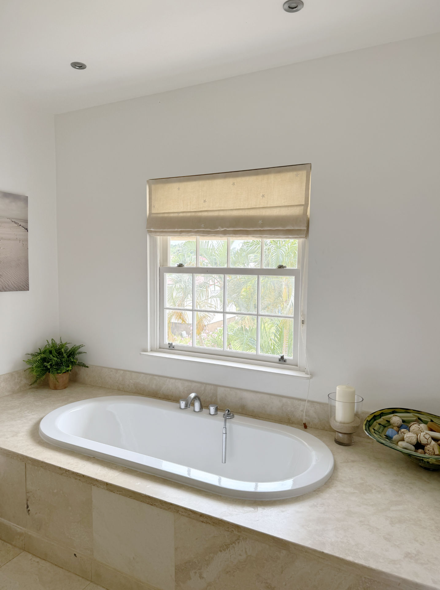 Bathroom with a built-in bathtub, a window with a beige Roman shade, a small green plant in a pot, candles and decorative shells on a tray.