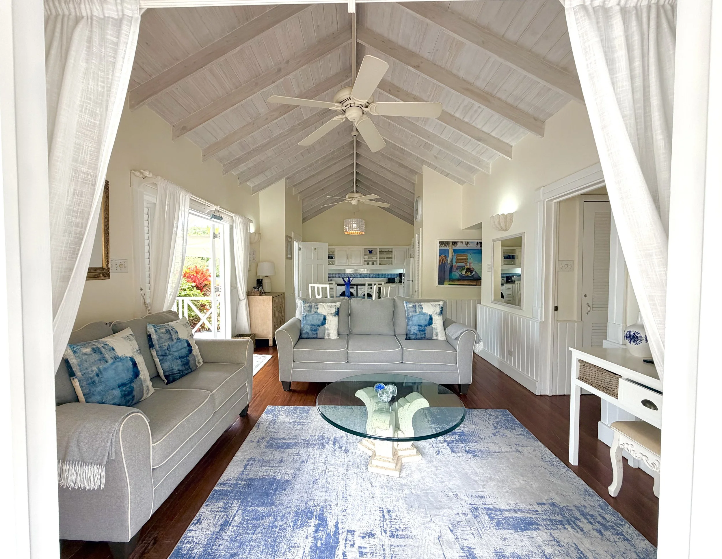 Living room with a vaulted wooden ceiling, ceiling fans, white curtains, a white couch with blue and white pillows, a glass coffee table, and tropical artwork on the walls.