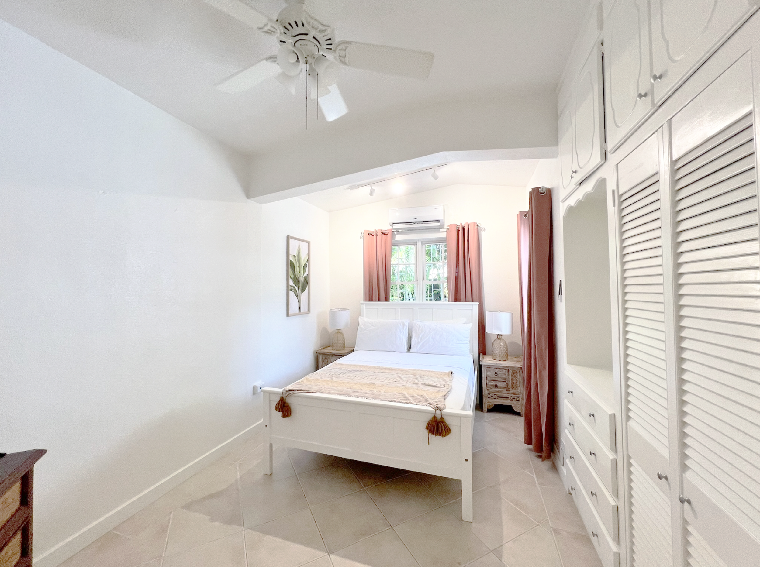 A bedroom with a white bed, pink curtains, a ceiling fan, and a window with a view of greenery.