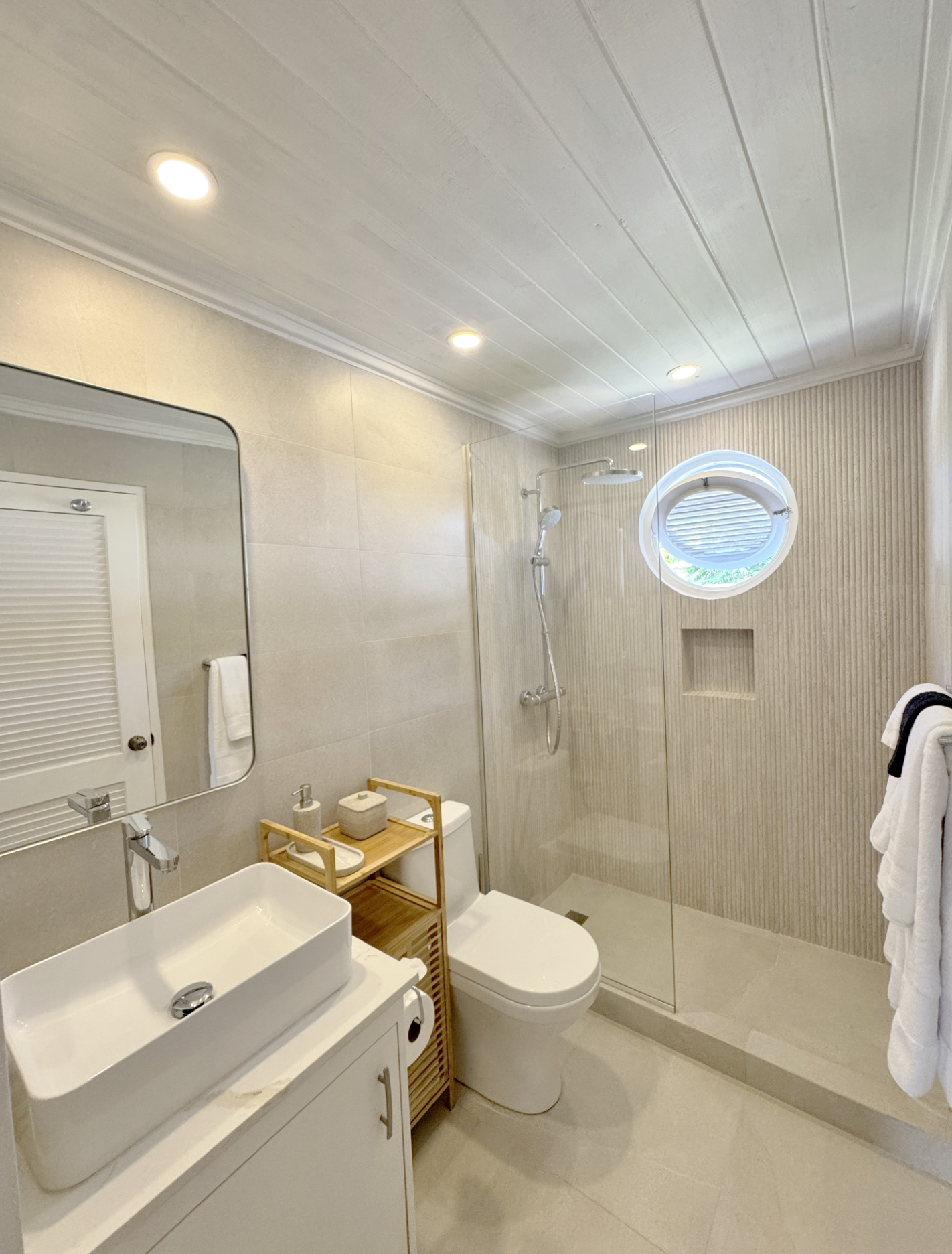 Modern bathroom with a walk-in shower, round window, vanity with rectangular sink, toilet, and beige tiled walls.