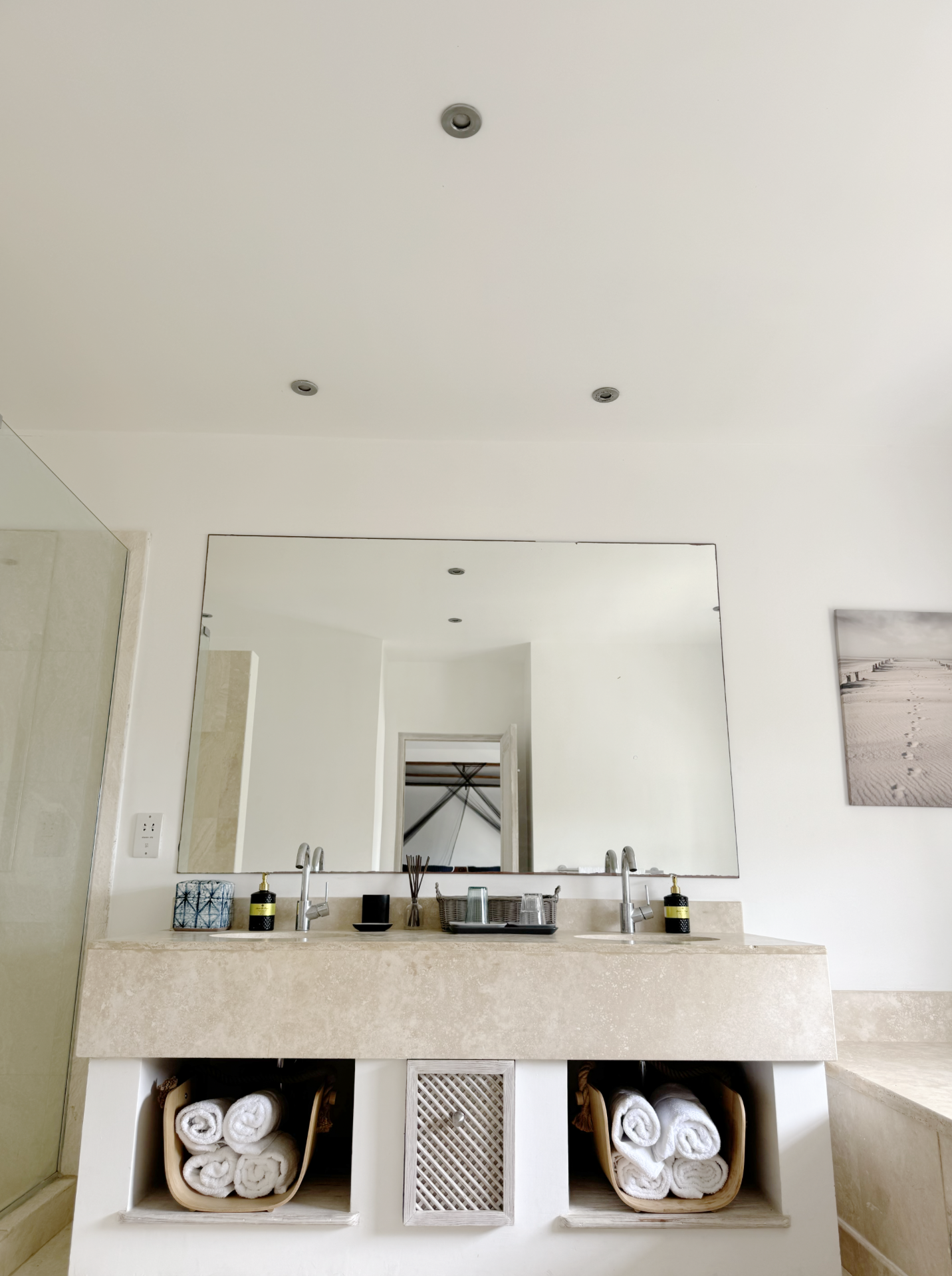 Bathroom vanity with a large mirror, two sinks, and storage shelves containing rolled white towels, soap dispensers, and glassware.