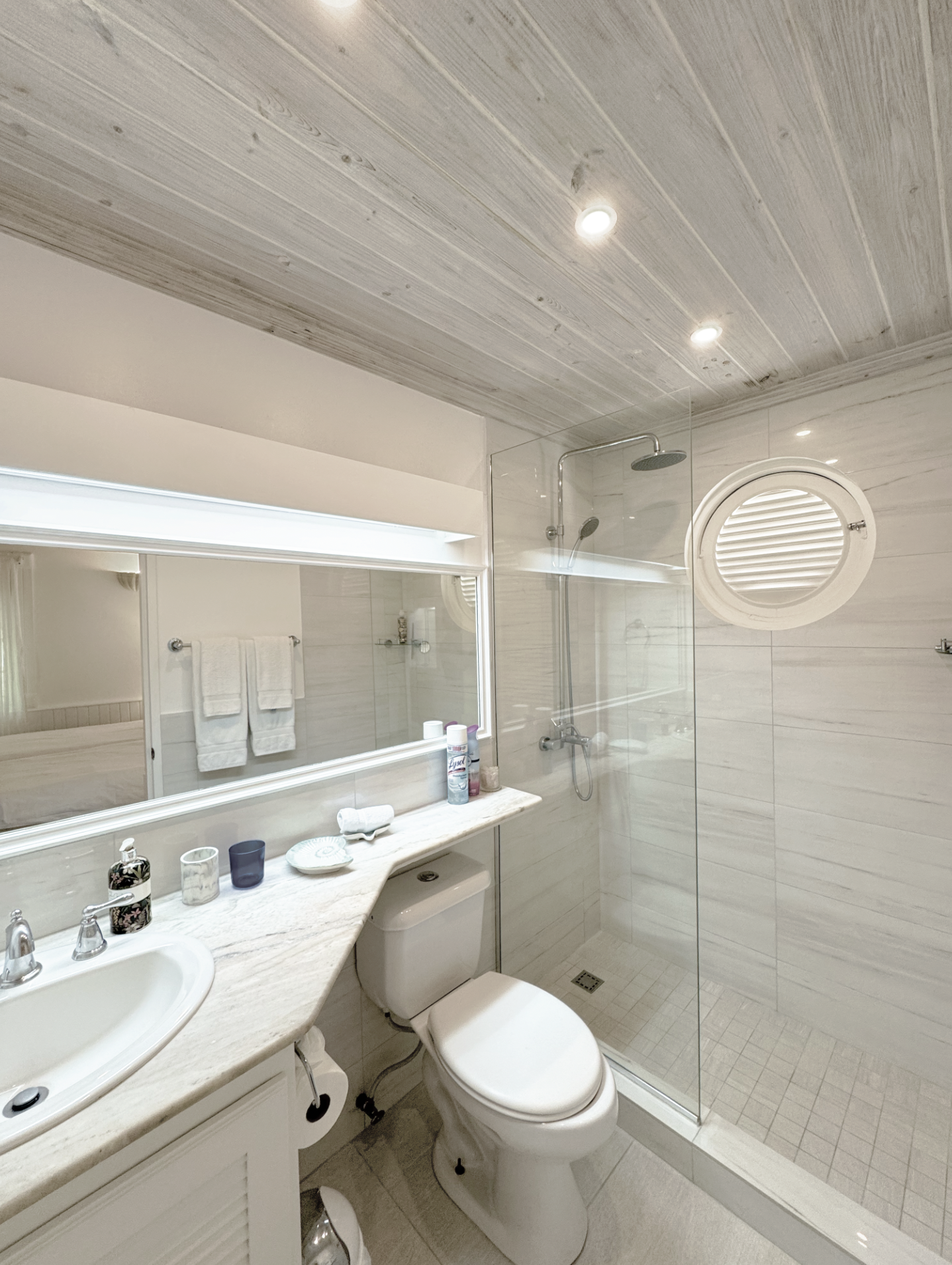 A modern bathroom with a beige marble countertop, a white sink, and a toilet. There is a large mirror above the counter, with toiletries and decorative items on it. A glass-enclosed shower with a rain showerhead and a round window on the tiled wall a