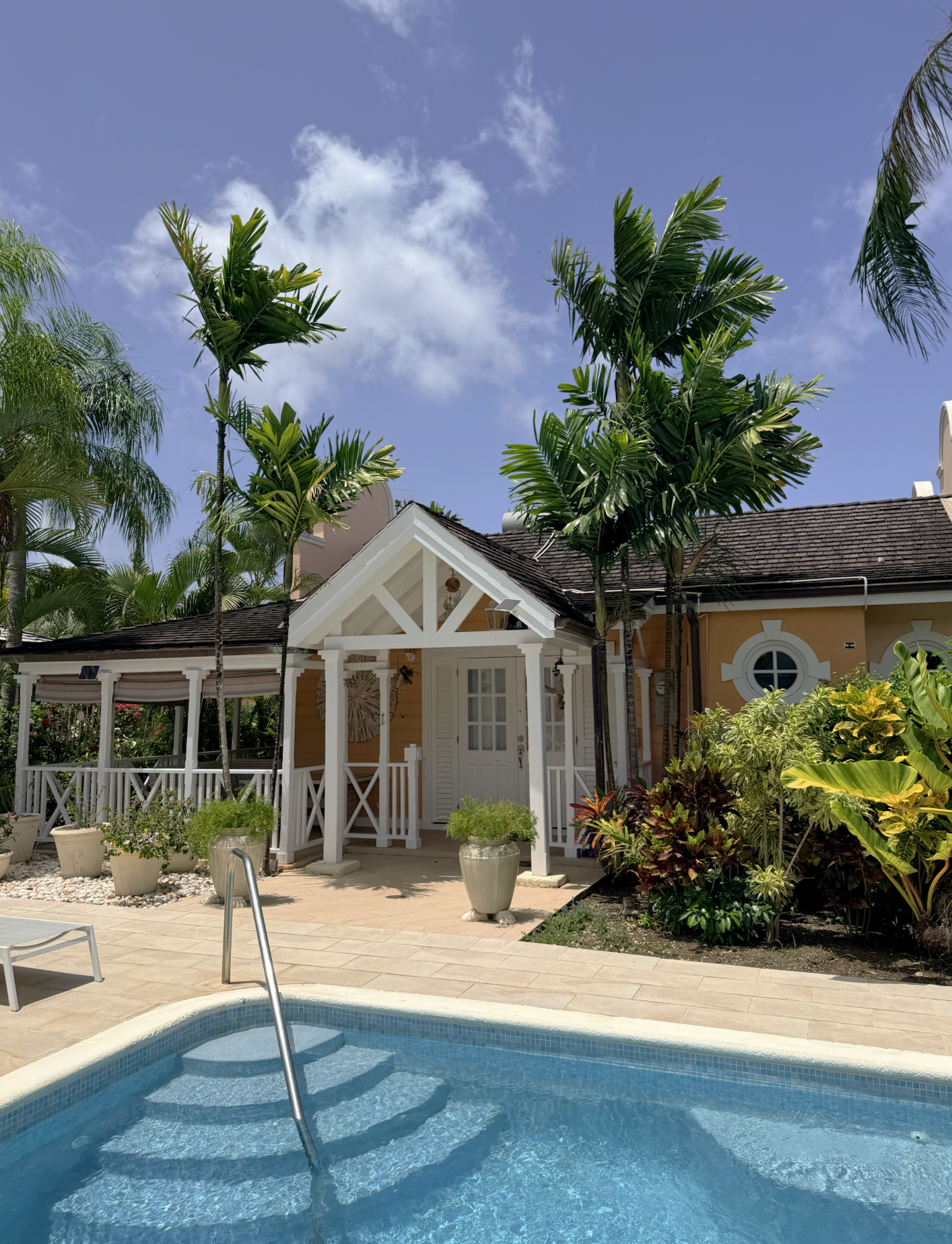 A house with a tan exterior and white trim, surrounded by palm trees and lush greenery. There is a swimming pool with a metal ladder in the foreground, and the sky is blue with scattered clouds.