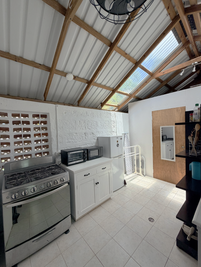 Kitchen area with a white refrigerator, microwave and toaster oven on a white cabinet, a gas stove, black shelving with bottles and containers, and a wooden door. The ceiling has exposed wooden beams and vent panels.