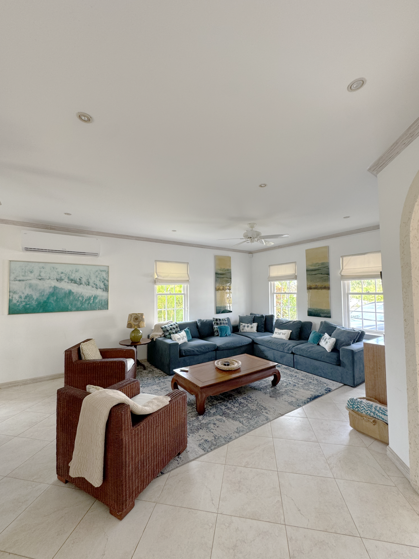 Living room with a large blue sectional sofa, two armchairs, a wooden coffee table, and artwork on the walls. Windows with blinds let in natural light.