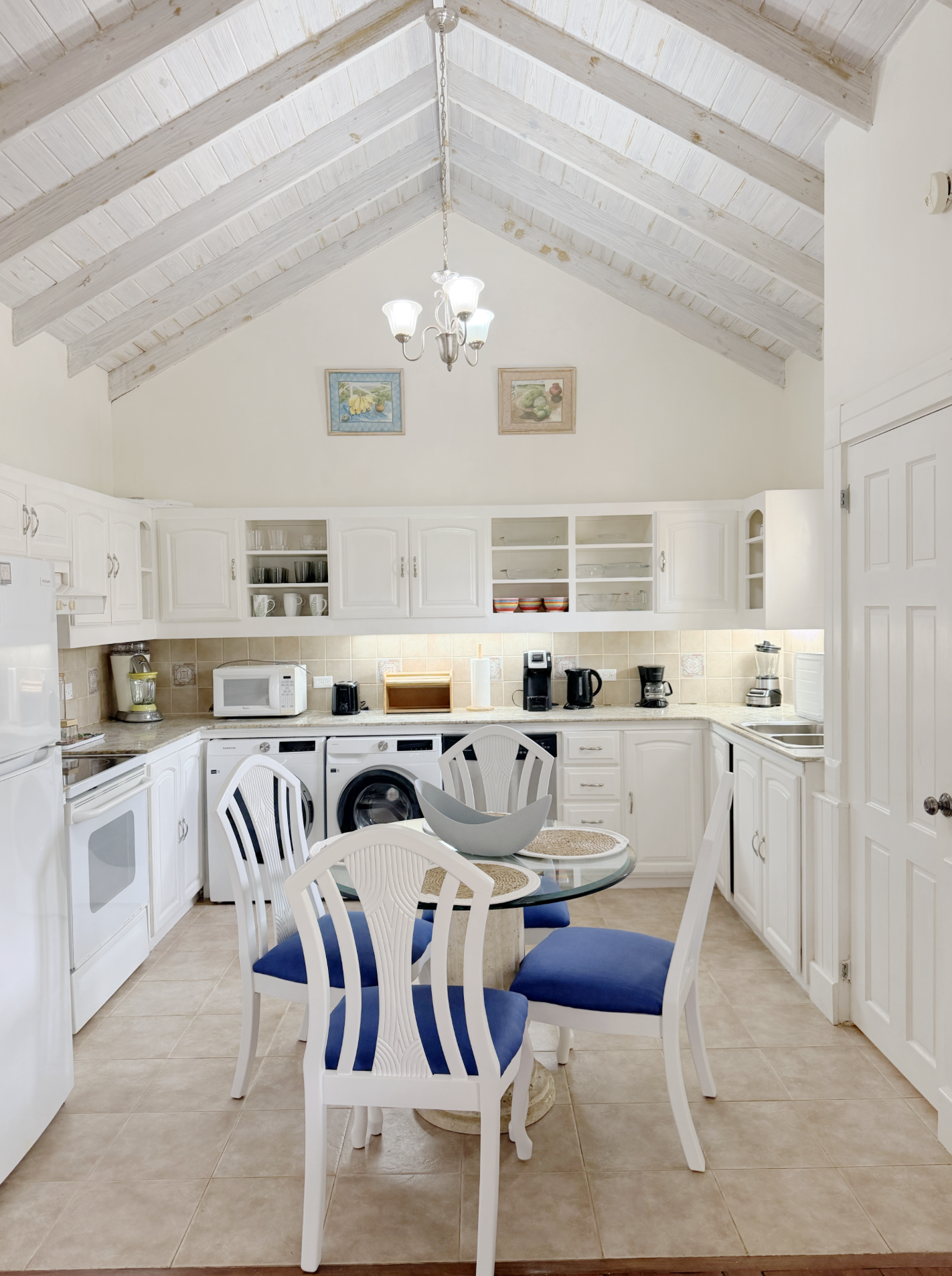 Kitchen with white cabinets, beige tiled floor, dining table with six chairs with blue seats, overhead chandelier, and various appliances on the countertop, including a microwave, coffee makers, and a blender.