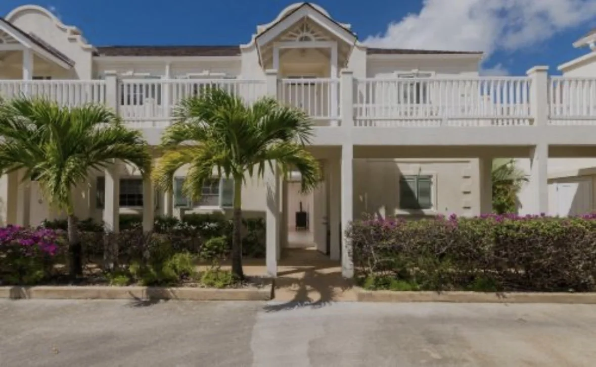 View of a white multi-story house with a front porch, balcony, palm trees, and flowering bushes under a blue sky.