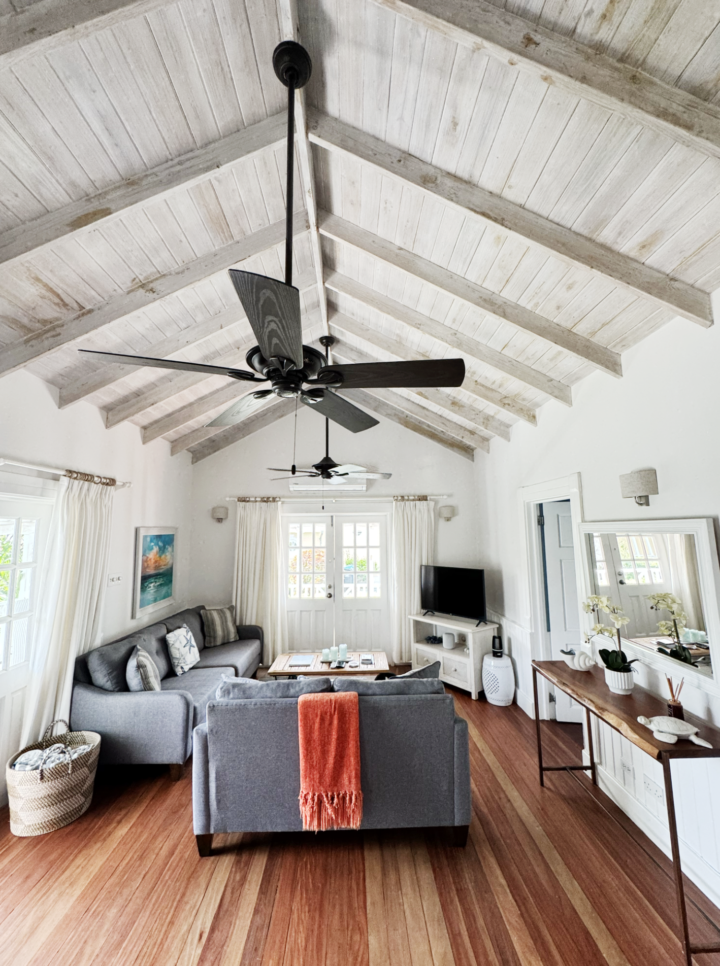 Living room with a vaulted, white-washed wooden ceiling featuring black ceiling fans. There is a gray sectional sofa with pillows, a wooden coffee table, and a white TV stand with a flat-screen TV. The room is bright with windows and a glass door, ad