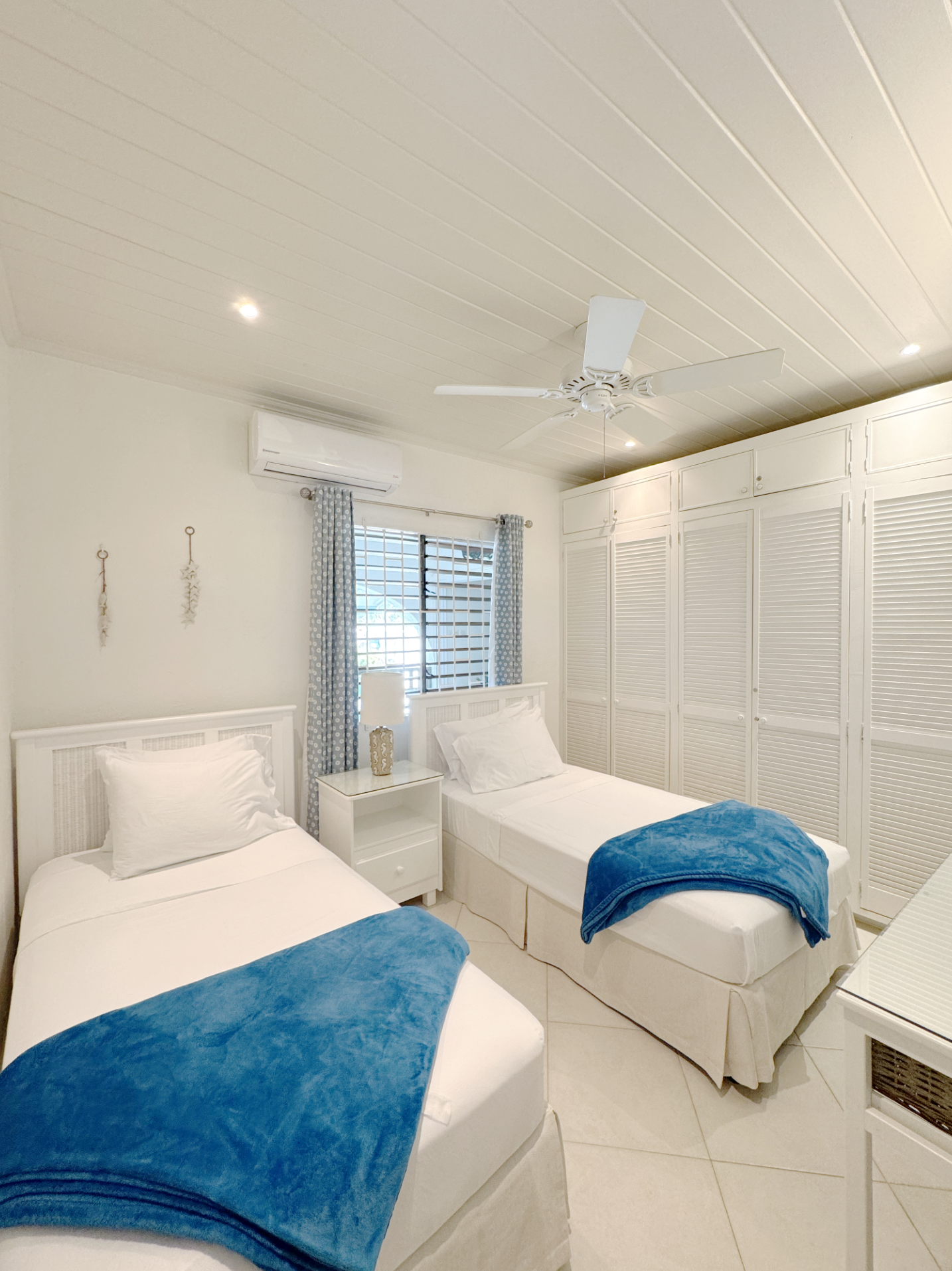 A bedroom with two single beds, each with white bedding and blue throws at the foot. Between the beds is a white nightstand with a decorative lamp. There are light-colored walls, a window with blue and white polka dot curtains, and a ceiling fan abov
