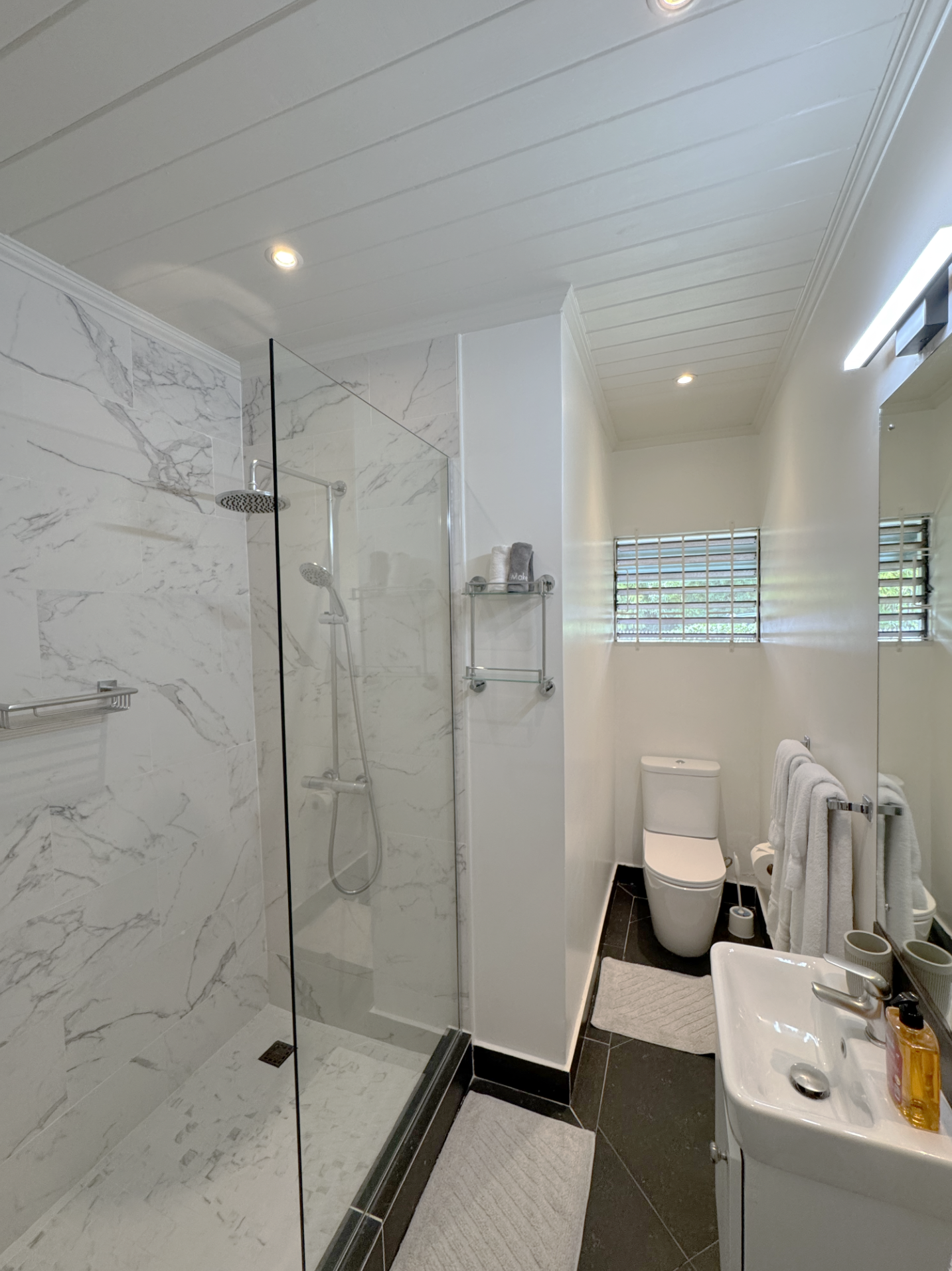 Bathroom with walk-in shower featuring marble tiles, glass partition, and ceiling-mounted rain shower head. White toilet, small white sink, mirror with light, and towel racks with towels. Small window with horizontal blinds, black tiled floor, and a 