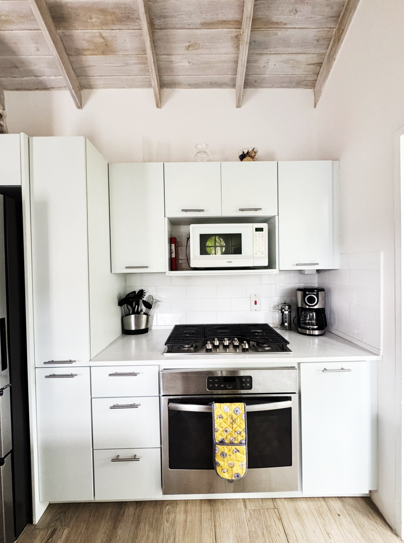 A modern kitchen with white cabinets, a stainless steel oven, a gas stove, a microwave above the stove, a coffee maker, utensils in a container, a fire extinguisher, and a yellow oven mitt hanging on the oven handle. The ceiling is made of wooden bea