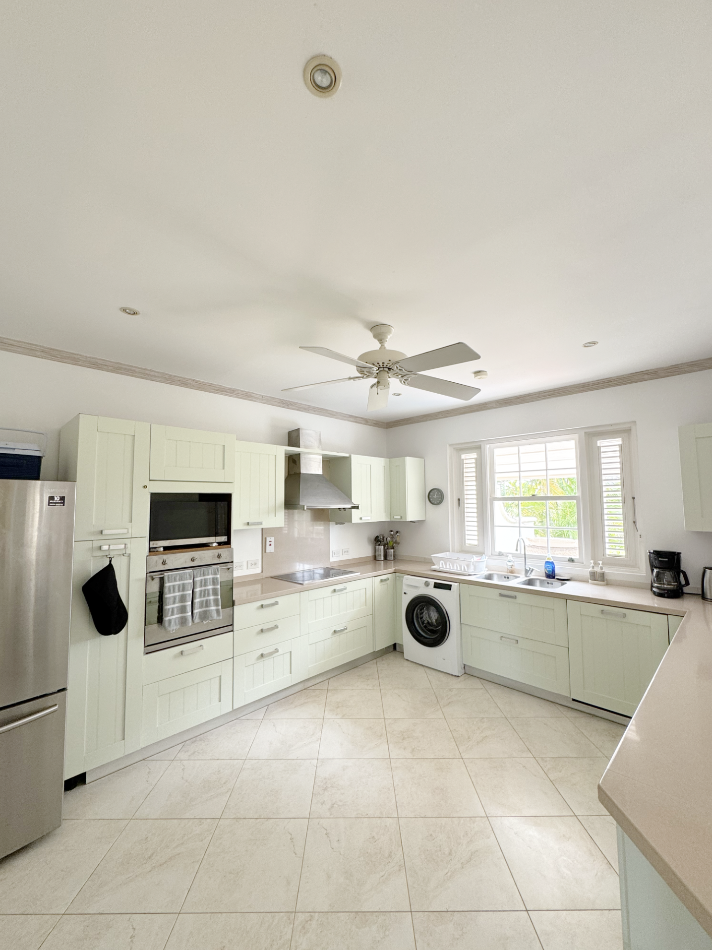 Bright kitchen with light green cabinets, white countertops, a window with shutters, and appliances including a refrigerator, microwave, oven, washing machine, coffee maker, and sink.