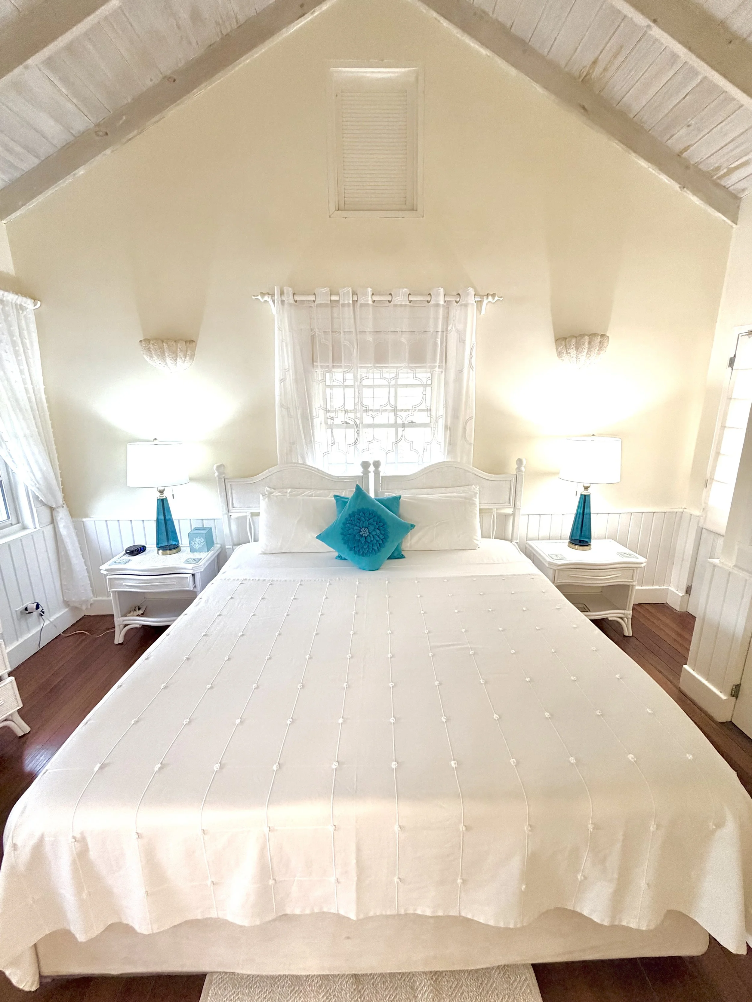 A bedroom with a white bed, teal pillows, two matching nightstands with lamps, and a window with white curtains. The ceiling is sloped with a vent in the center.