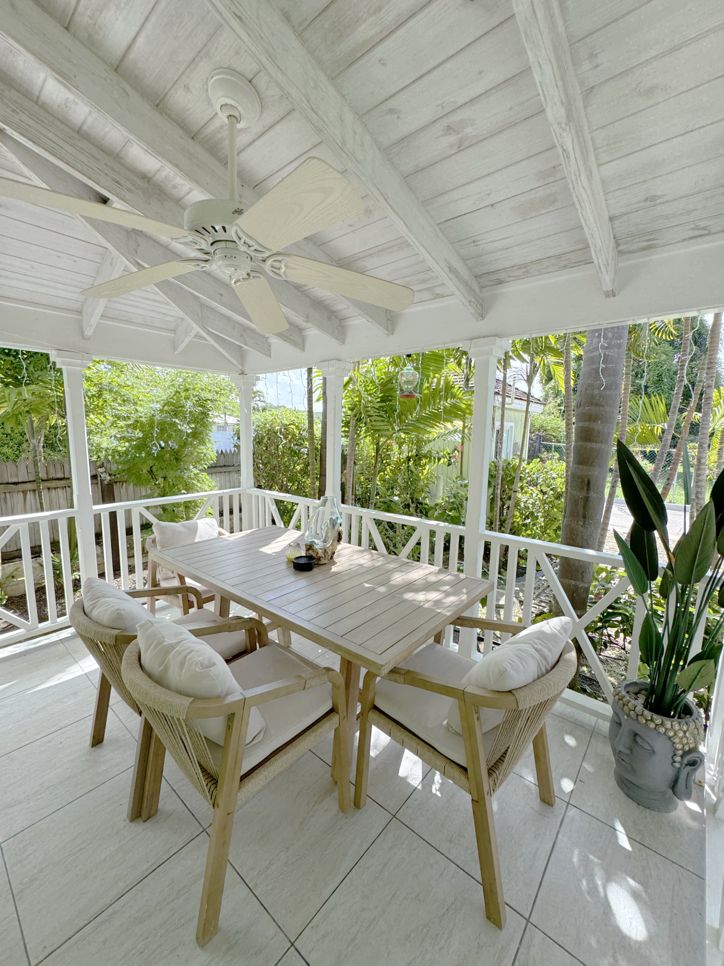 An outdoor patio area with a wooden dining table surrounded by cushioned chairs, a ceiling fan overhead, and lush green plants and trees in the background.