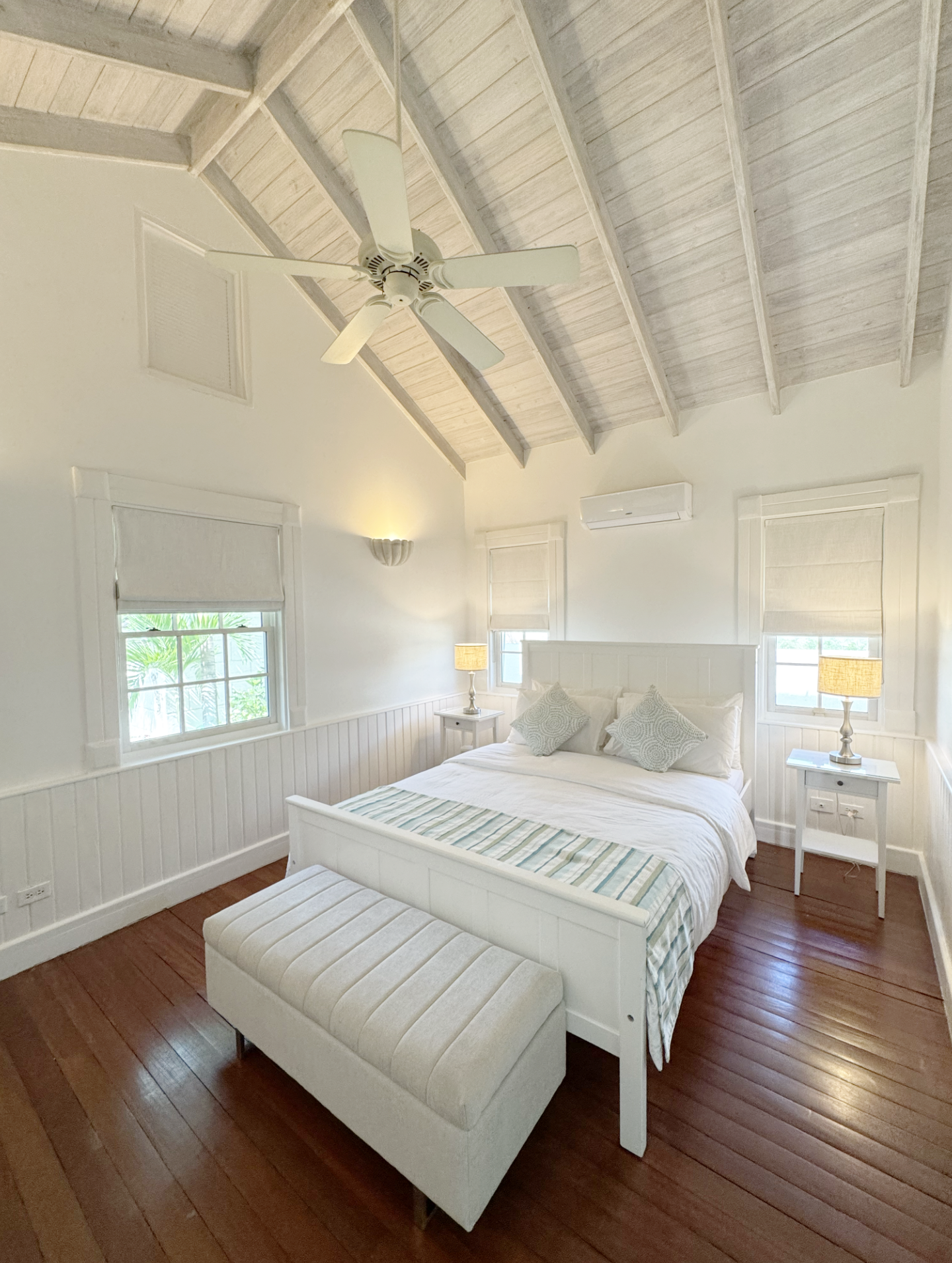 A cozy bedroom with a white wooden bed, striped bedding, and matching pillows. Two nightstands with lamps flank the bed, and three windows with light-colored blinds are on the walls. The room has a wooden floor, and a ceiling fan with light-colored b