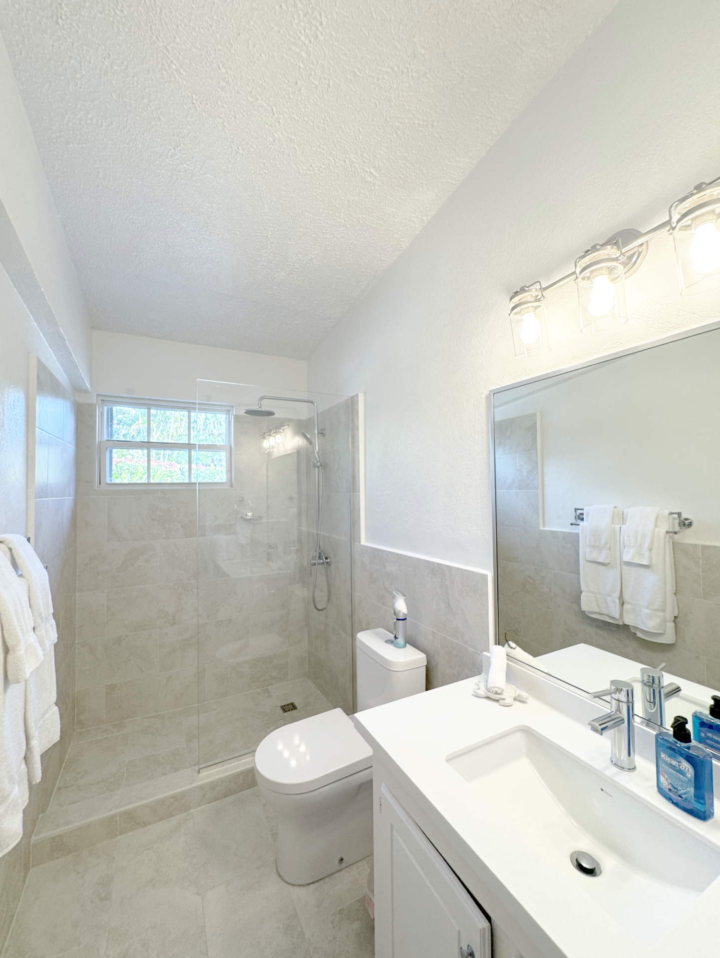 Modern bathroom with beige tiled flooring and walls, a walk-in shower with glass door, a window, a white toilet, a white vanity with a sink, a large mirror, and mounted light fixtures.