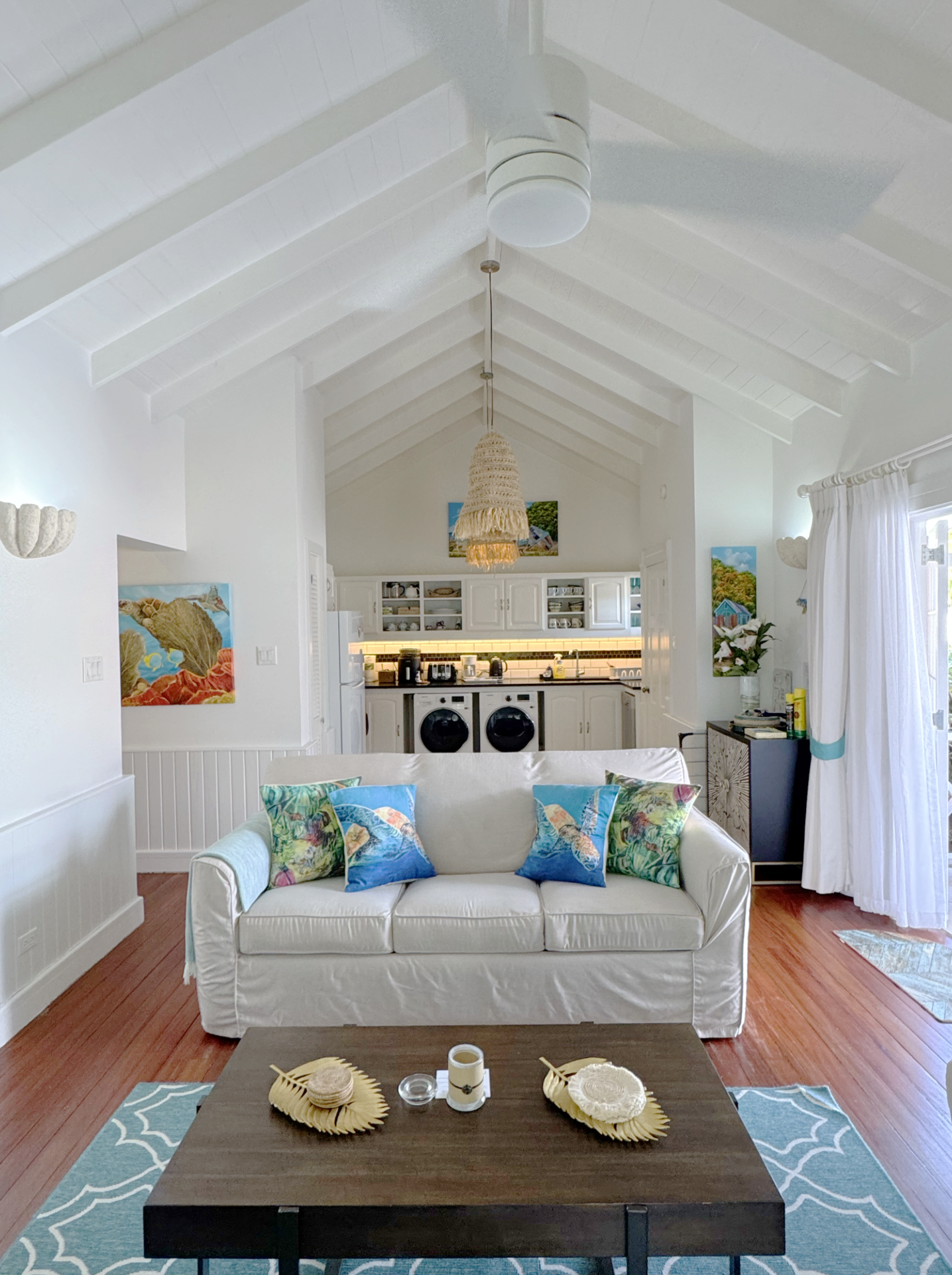 Living room with white sofa, colorful pillows, a dark wooden coffee table with decorative items, blue area rug, open kitchen with white cabinets, tiled backsplash, and hanging light fixtures, natural light from windows with white curtains.