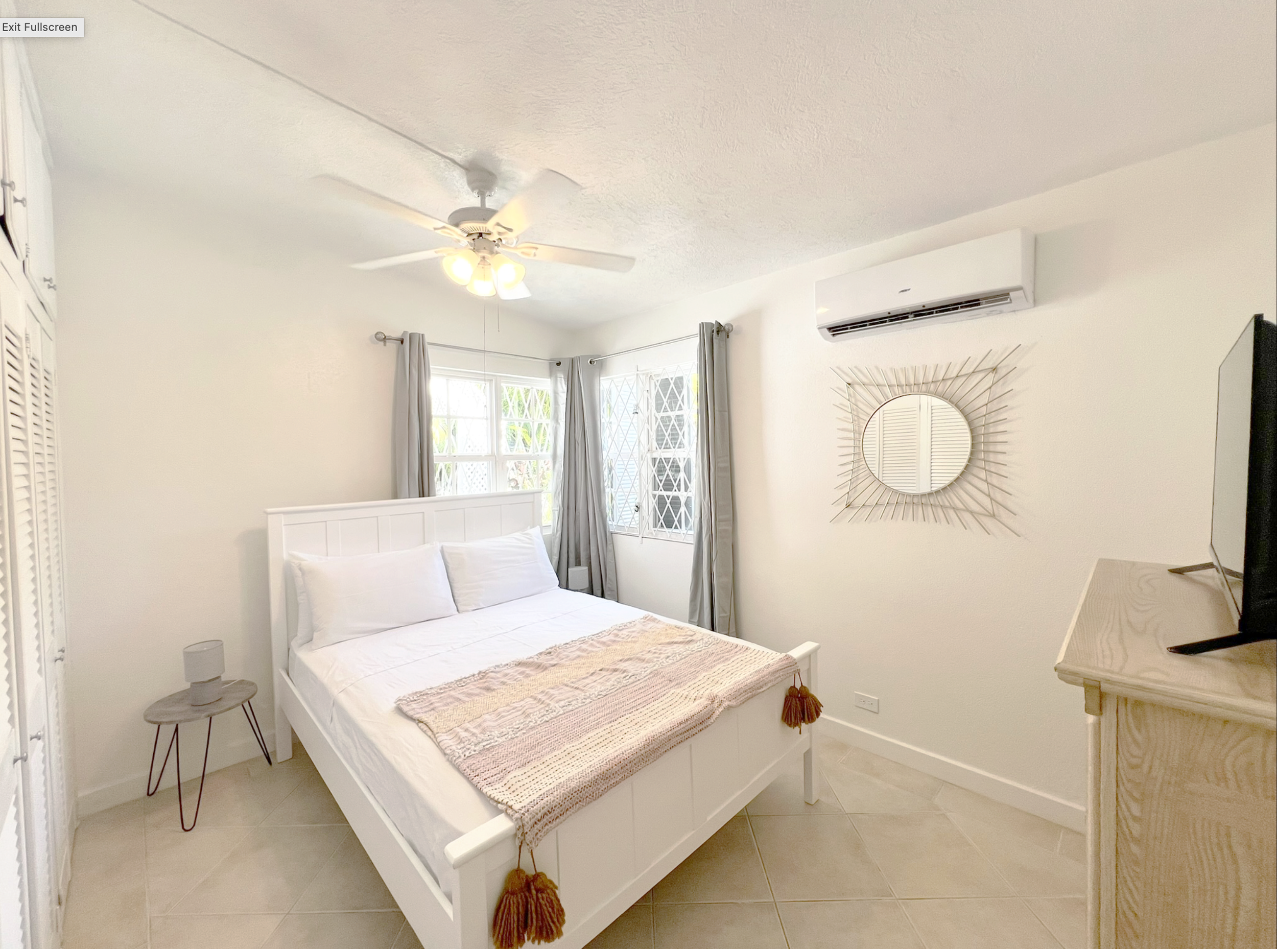 Bright bedroom with white bed, beige throw blanket, nightstand with lamp, window with curtains, wall mirror, and air conditioning unit.