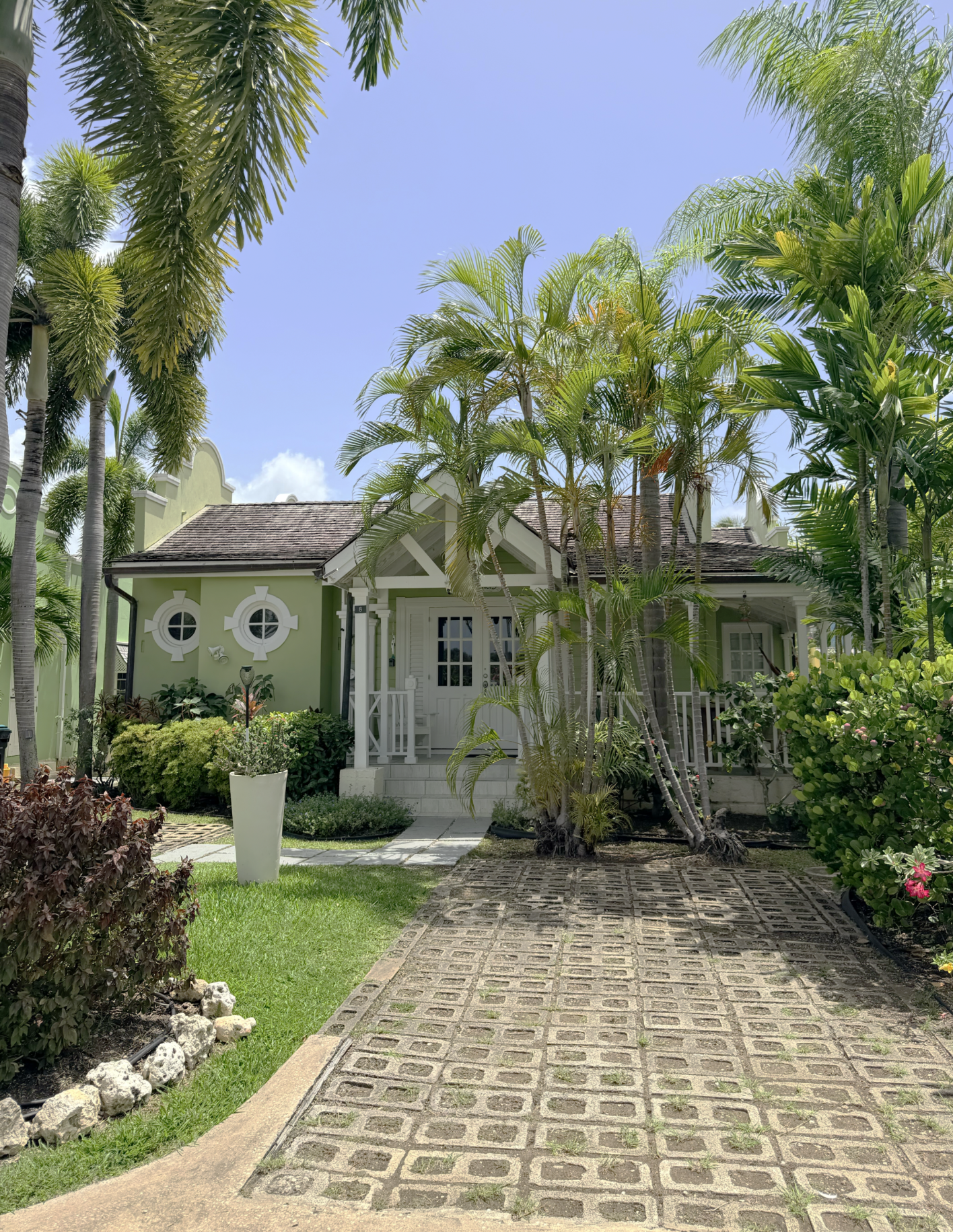 A small, green house with white trim and a shingled roof surrounded by a lush garden with various palm trees and flowering bushes, and a brick-paved driveway leading to the front door on a bright, sunny day.