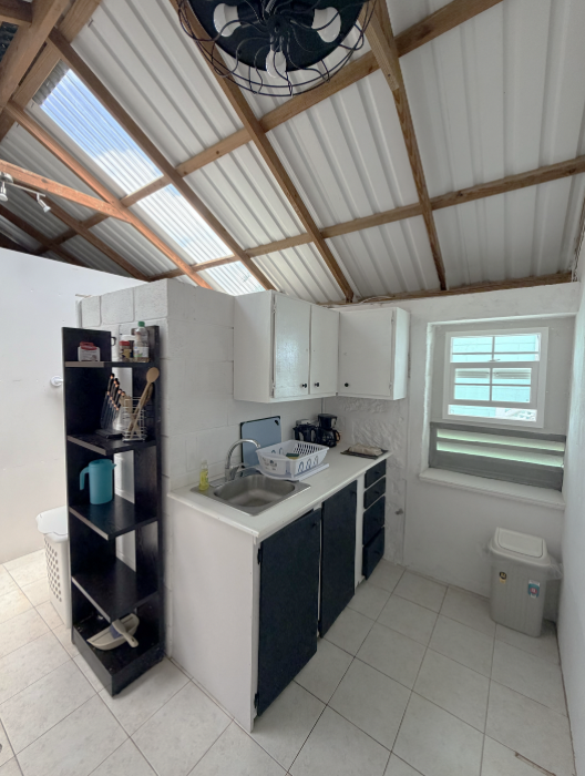 Small kitchen with a white tiled floor, white walls, and white cabinets. There is a window with closed blinds, a black open shelving unit, and a small sink with a dish rack. Ceiling has exposed wooden beams and a ceiling fan.