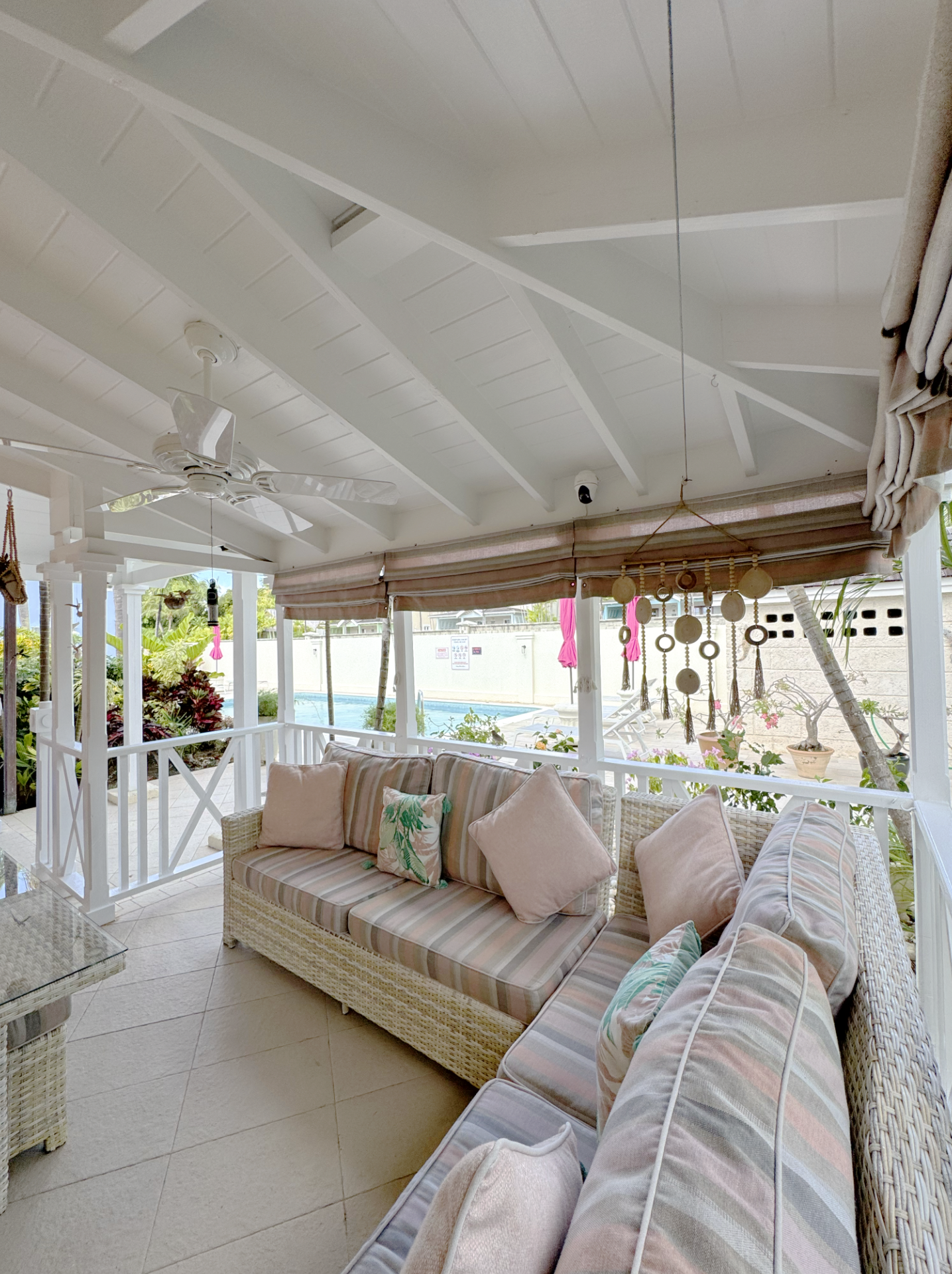 Outdoor porch seating area with wicker furniture, pastel cushions, ceiling fan, hanging decorations, and view of garden and pool area.