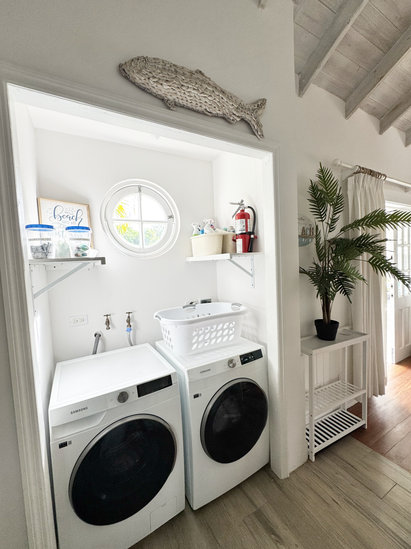 A laundry room with front-loading washing machine and dryer, laundry basket on top, some shelves with supplies, a fire extinguisher, a window, a plant, and a white shelf with a slatted lower shelf.