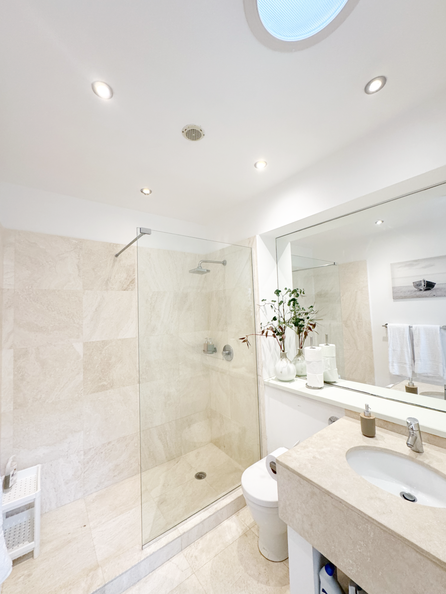Modern bathroom with beige tiled walls and floor, a glass shower enclosure with a rain showerhead, a large wall mirror, a white vanity with a sink, decorative plants, and bathroom accessories.
