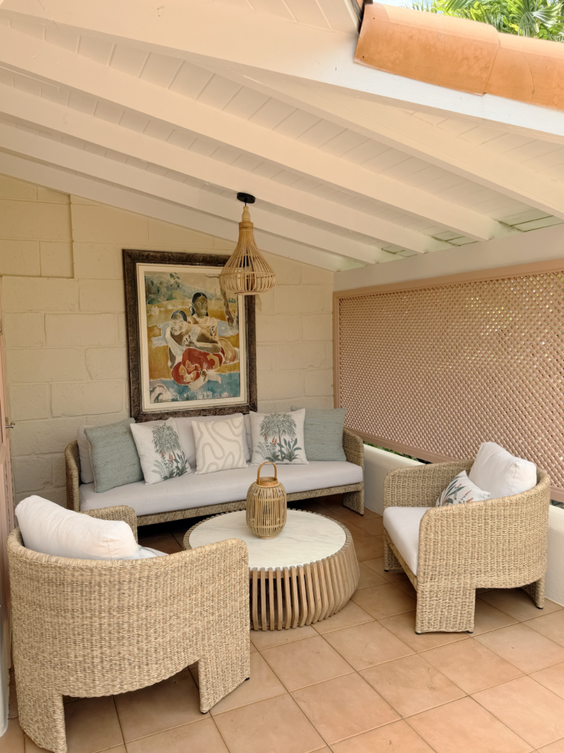 A cozy outdoor seating area with a white couch and two wicker chairs arranged around a round coffee table with a decorative vase. There is a framed painting of children on the wall behind the couch, and a pink lattice screen on one side. The ceiling 