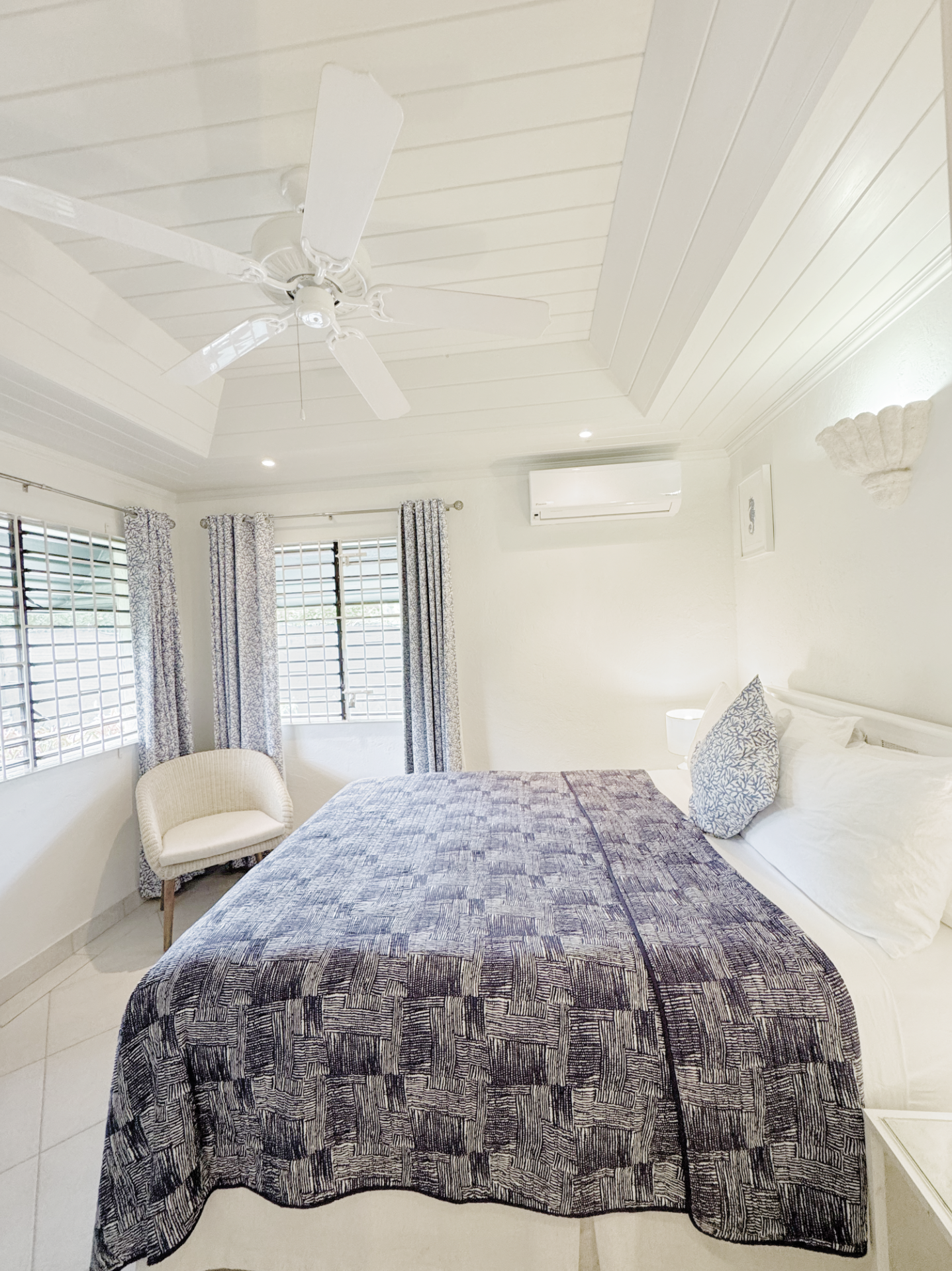 A bedroom with a bed covered in a dark patterned quilt, two pillows with matching pillowcases, a beige armchair, and windows with curtains. The ceiling has a white fan and recessed lighting, and there's an air conditioning unit on the wall.